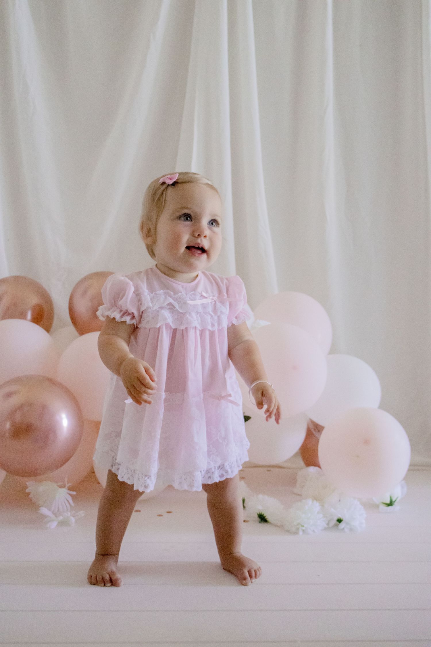 Smiling baby in lace pink dress standing against white backdrop with pastel balloons.