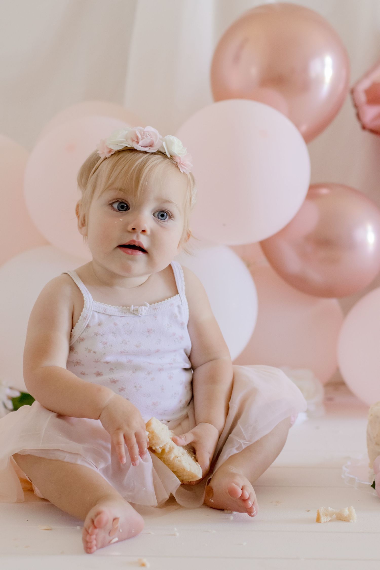 Baby in white dress with floral headpiece sitting with snack against pink balloons.