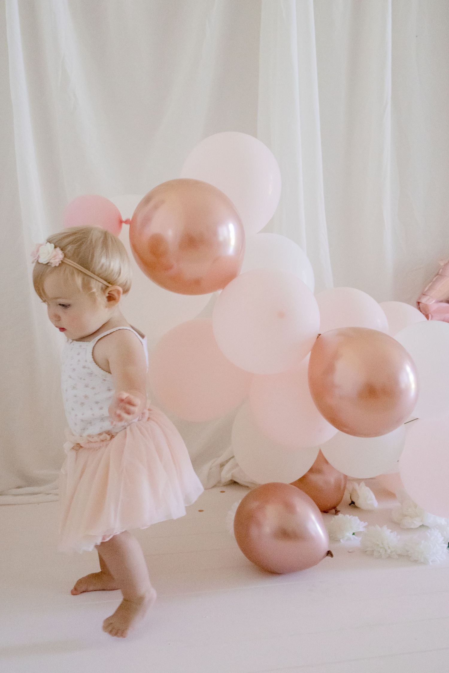 Small child in tulle dress walking past decorative balloon arrangement against white backdrop.