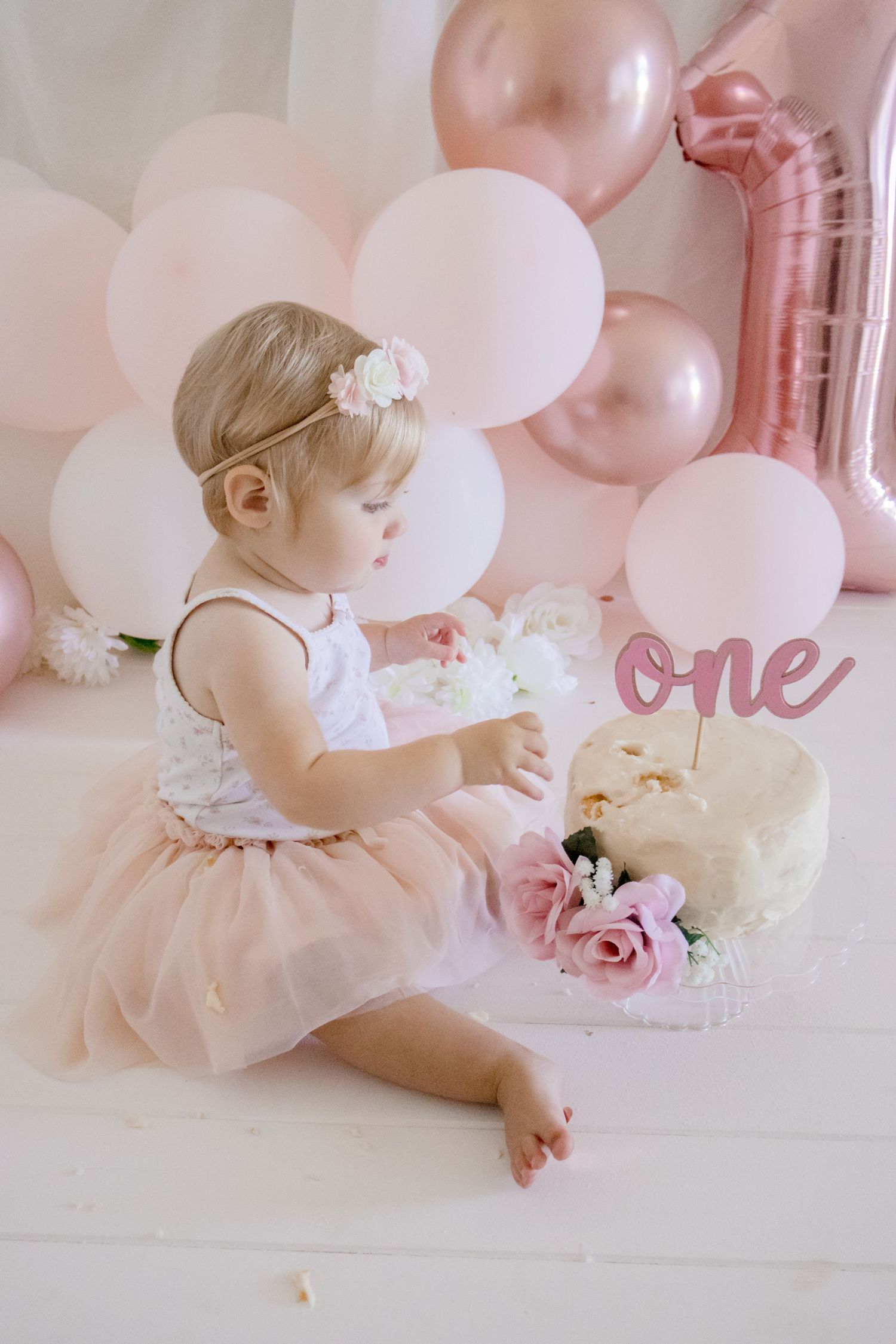 Profile shot of baby exploring birthday setup with pink balloons and white cake.