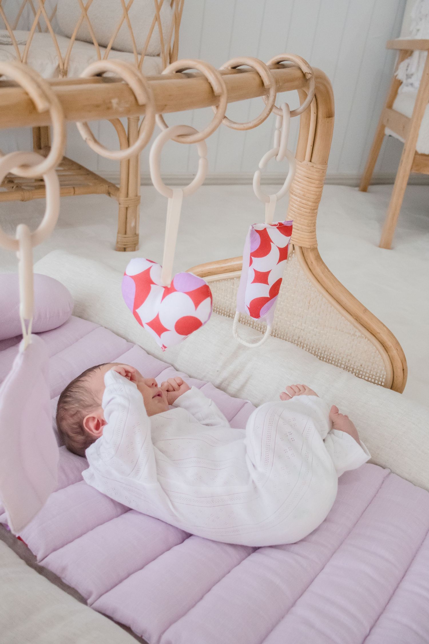 A baby lies on a quilted purple mat beneath wooden play rings with red and white patterned toys hanging above.