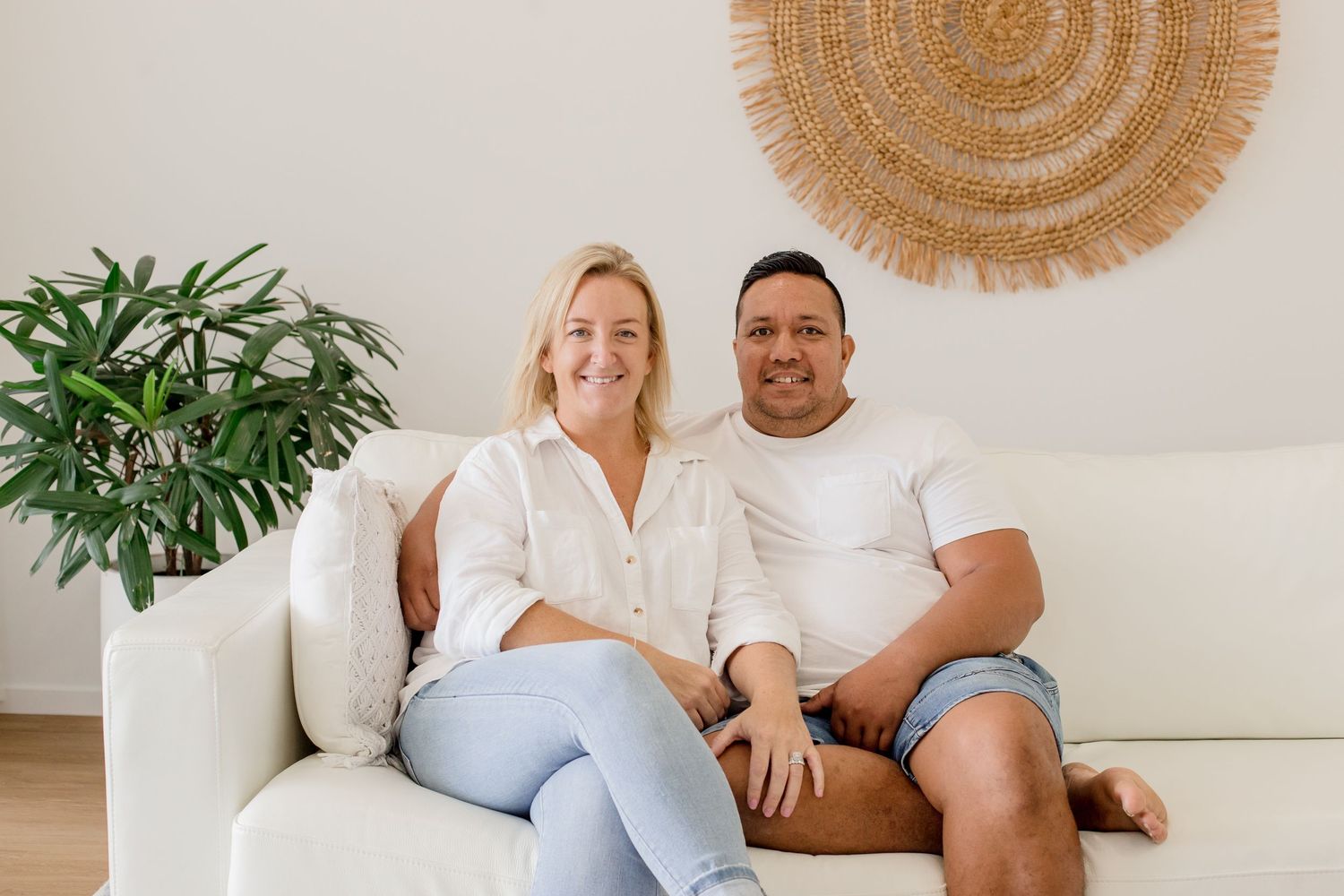 Happy couple relaxes on white sofa beneath natural woven wall decor.