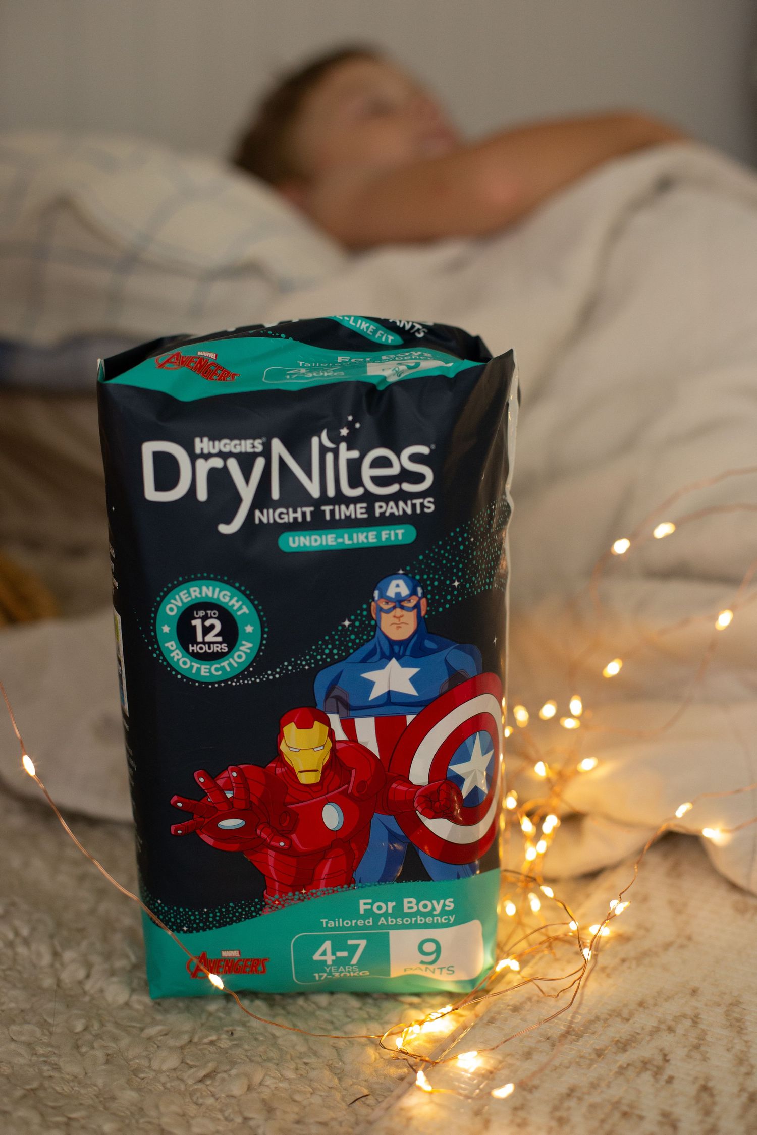 Package of Huggies DryNites nighttime pants with superhero design next to string lights.