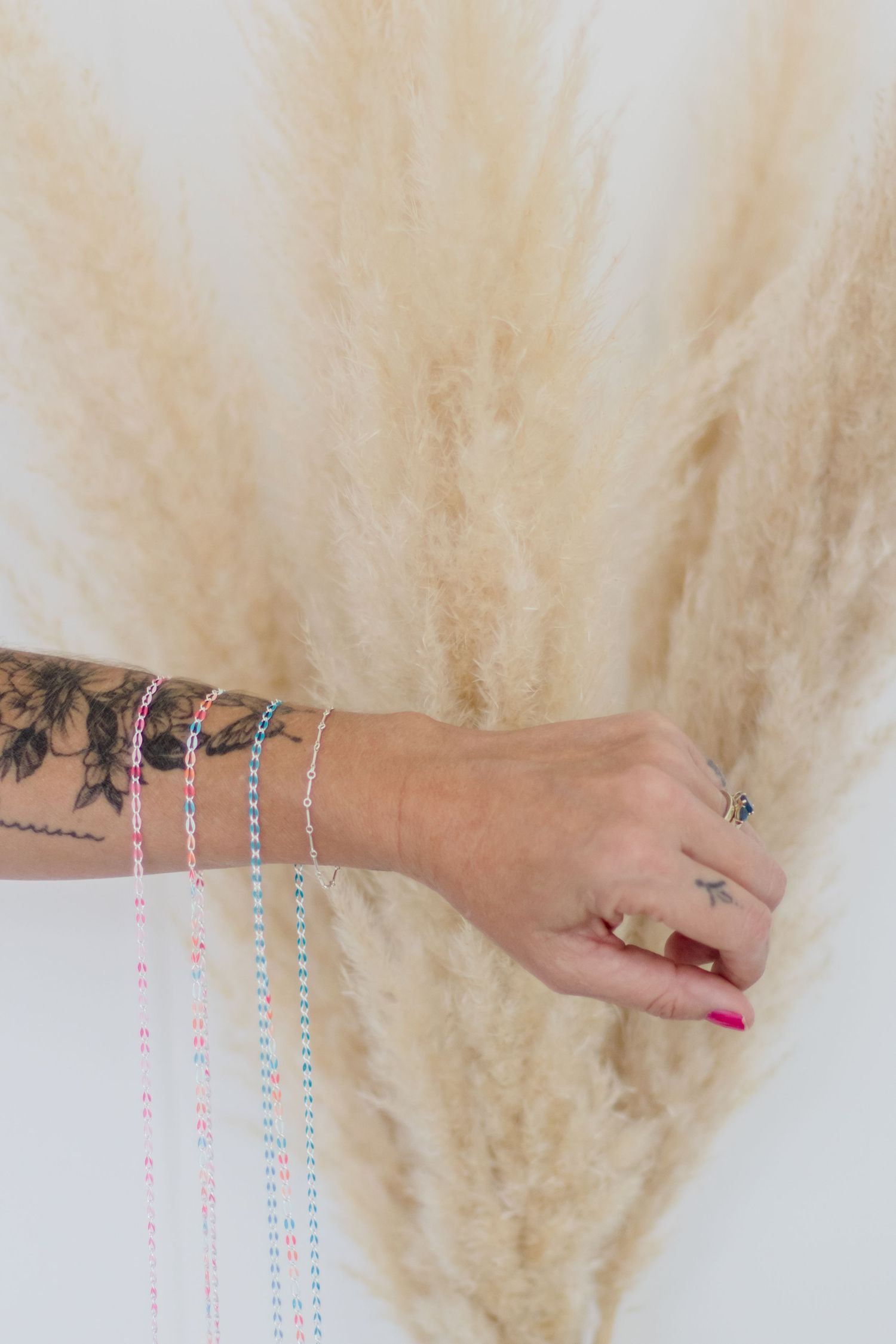 Tattooed arm wearing delicate pastel beaded bracelets against a soft beige pampas grass background.