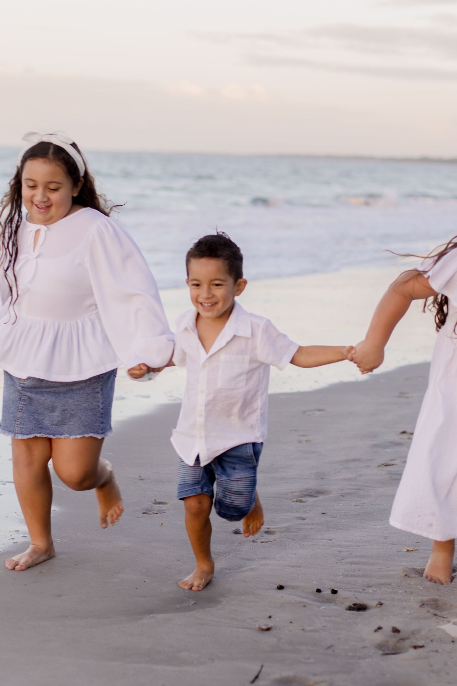 Children in white outfits run and play together joyfully on a beach at sunset along the ocean shore.