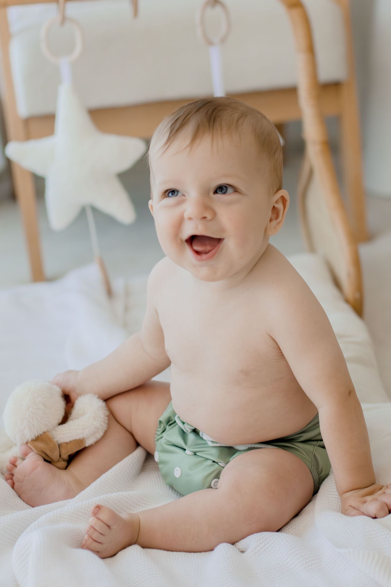 Happy baby sitting up wearing a green cloth diaper, smiling brightly in a bright nursery with star toys.