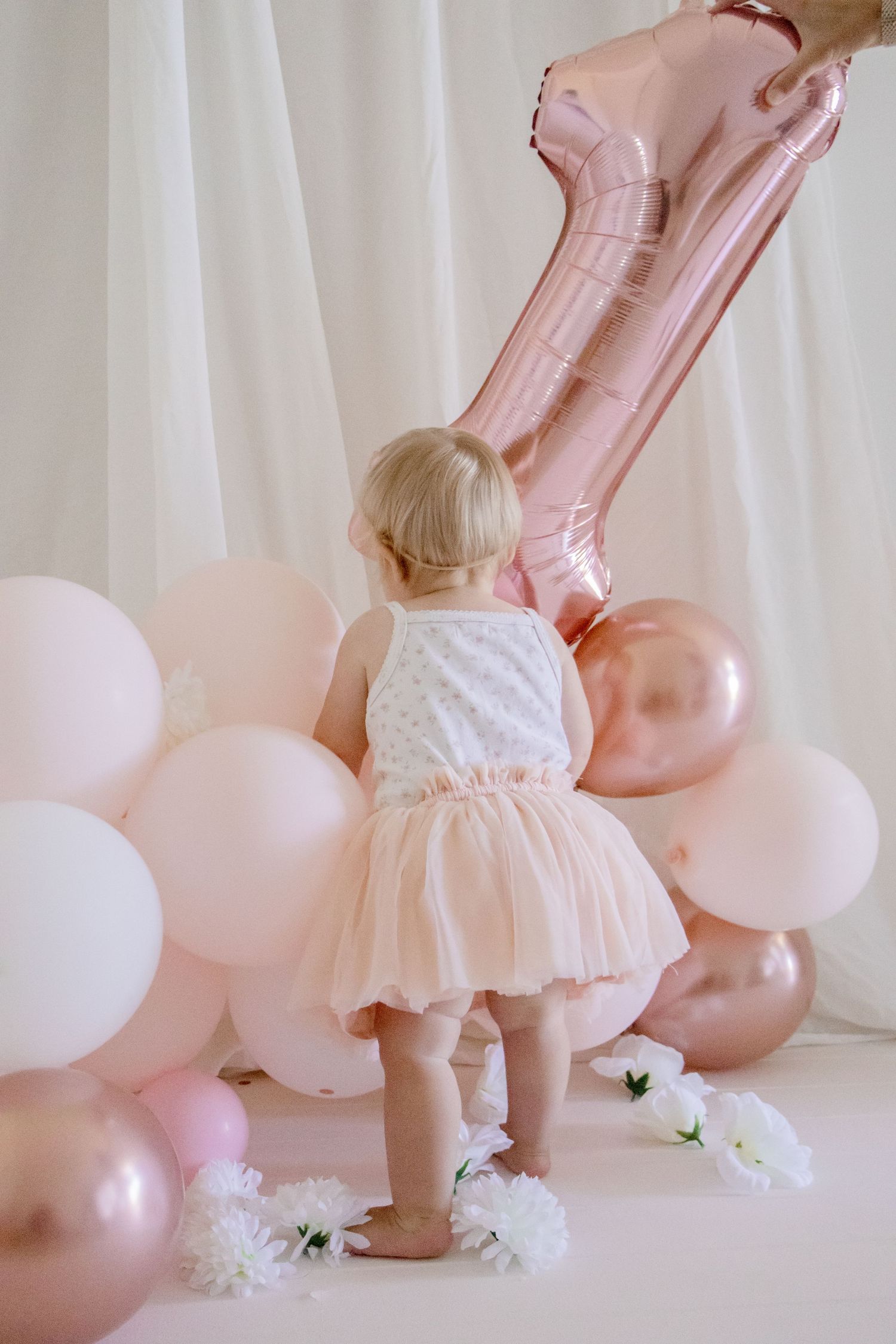 A toddler in a tulle dress stands near a large rose gold number one balloon and pink decorations.