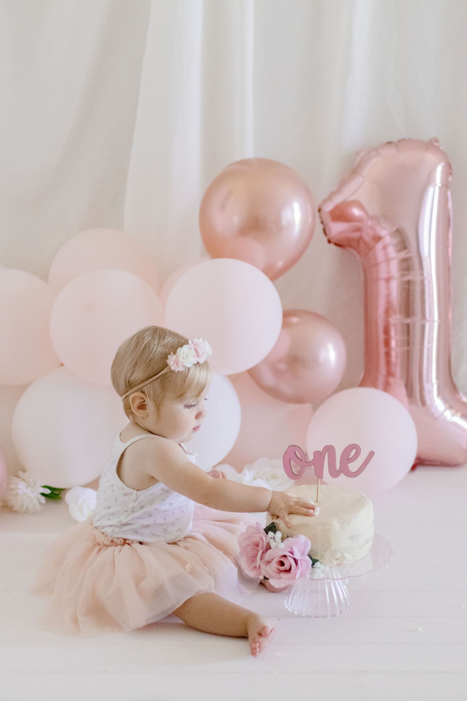 Elegant first birthday photoshoot with rose gold balloons and white cake decorations.