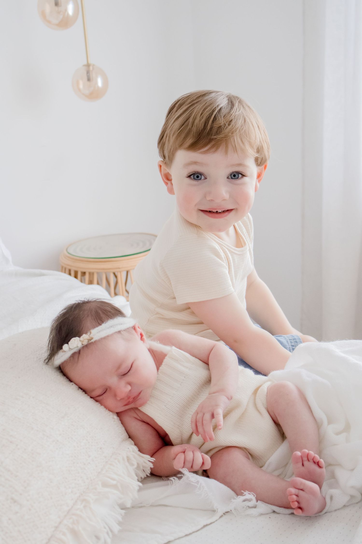 Siblings posing together on white bed with soft lighting and rattan decor.