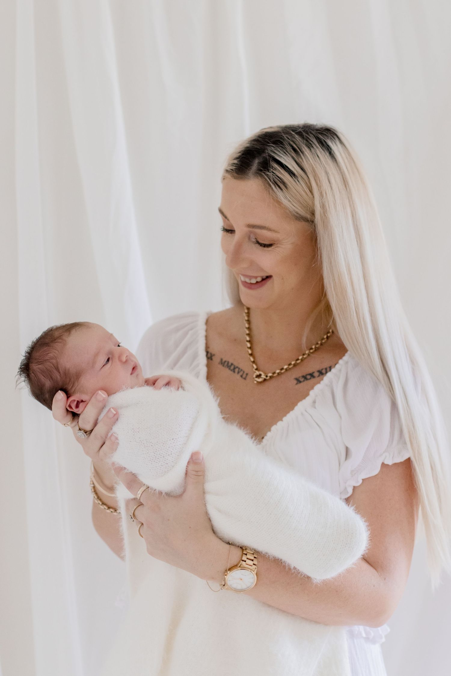 Portrait of newborn being held in white wrap while wearing gold jewelry.