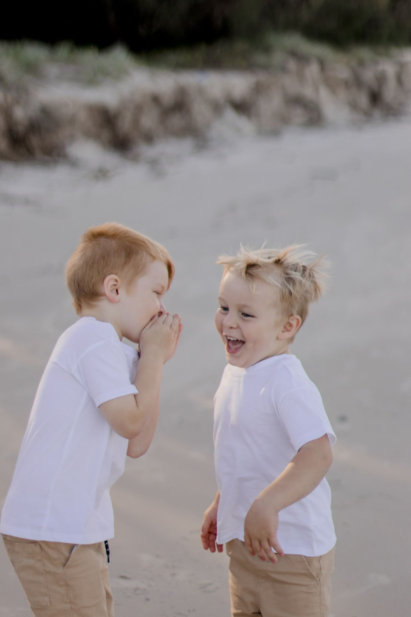 Two small children in white shirts share secrets and laugh on the beach.
