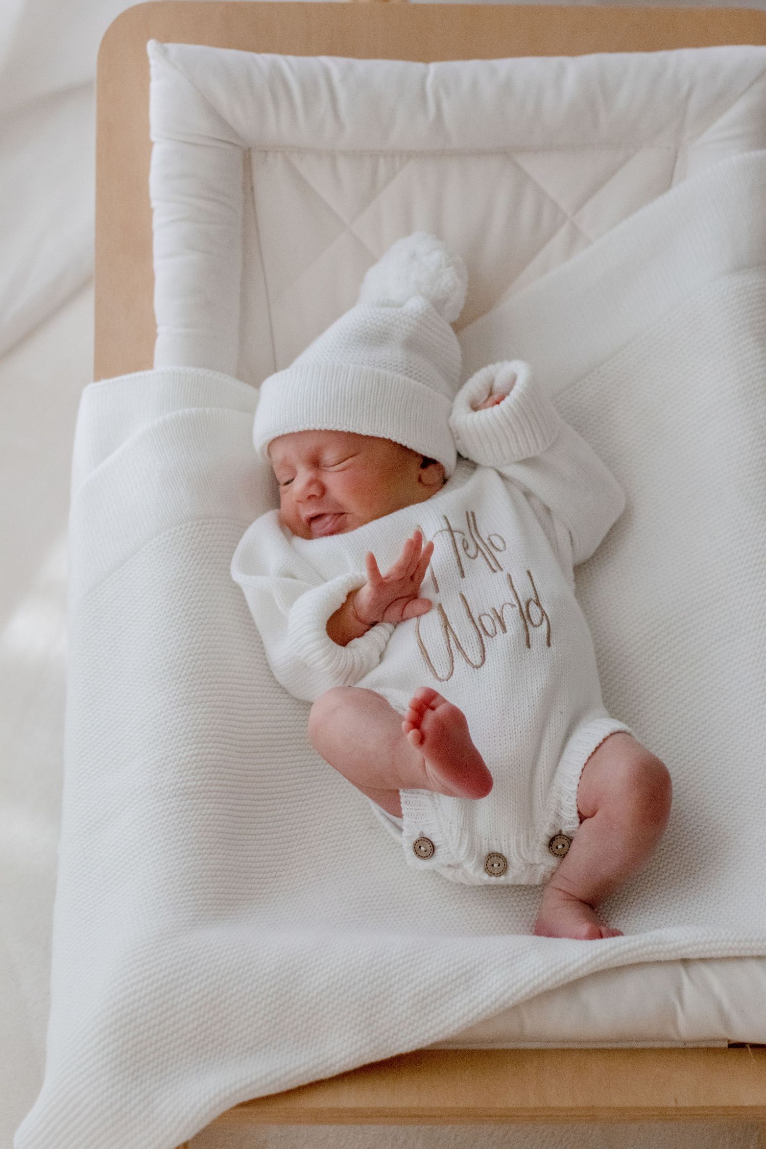Sleeping newborn in white knit outfit and beanie rests peacefully on plush white bedding.