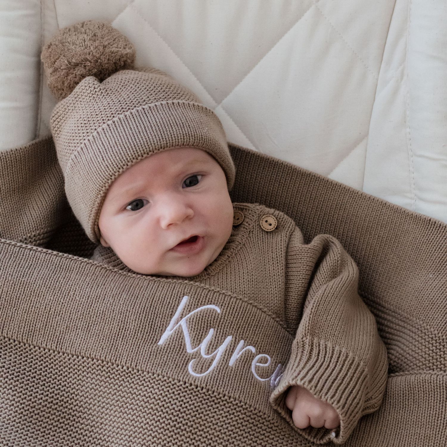 Baby in brown knit sweater and pink hat resting on textured brown cushion with embroidered text.