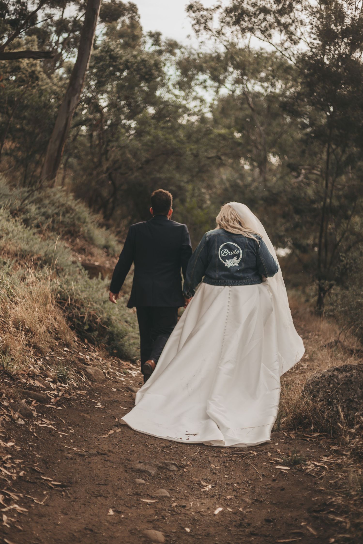 A couple sits together on a dirt trail surrounded by trees while wearing a wedding dress and black jacket.