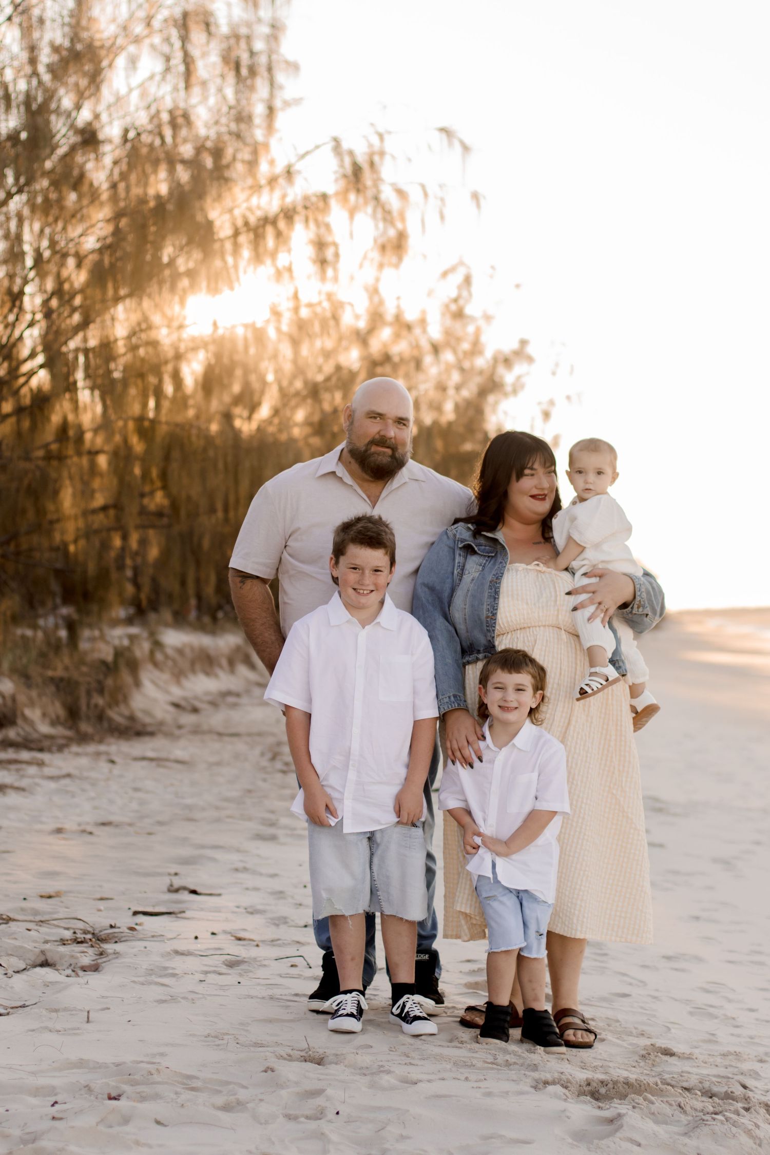 Family wearing coordinated white and neutral outfits poses together on a sandy beach at sunset creating a dreamy mood.