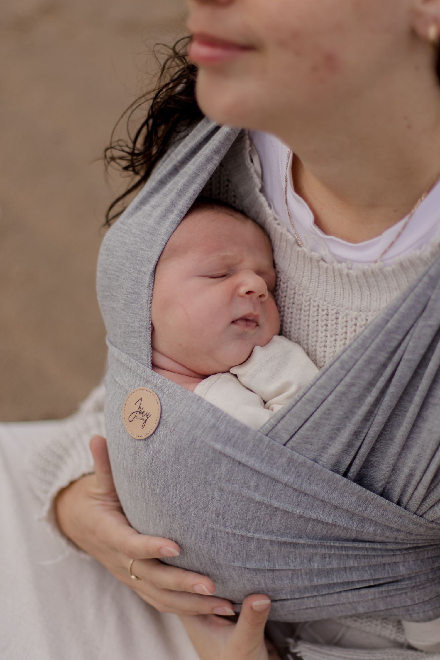 Peaceful newborn baby sleeping in soft grey fabric baby carrier against parent wearing white knit sweater.