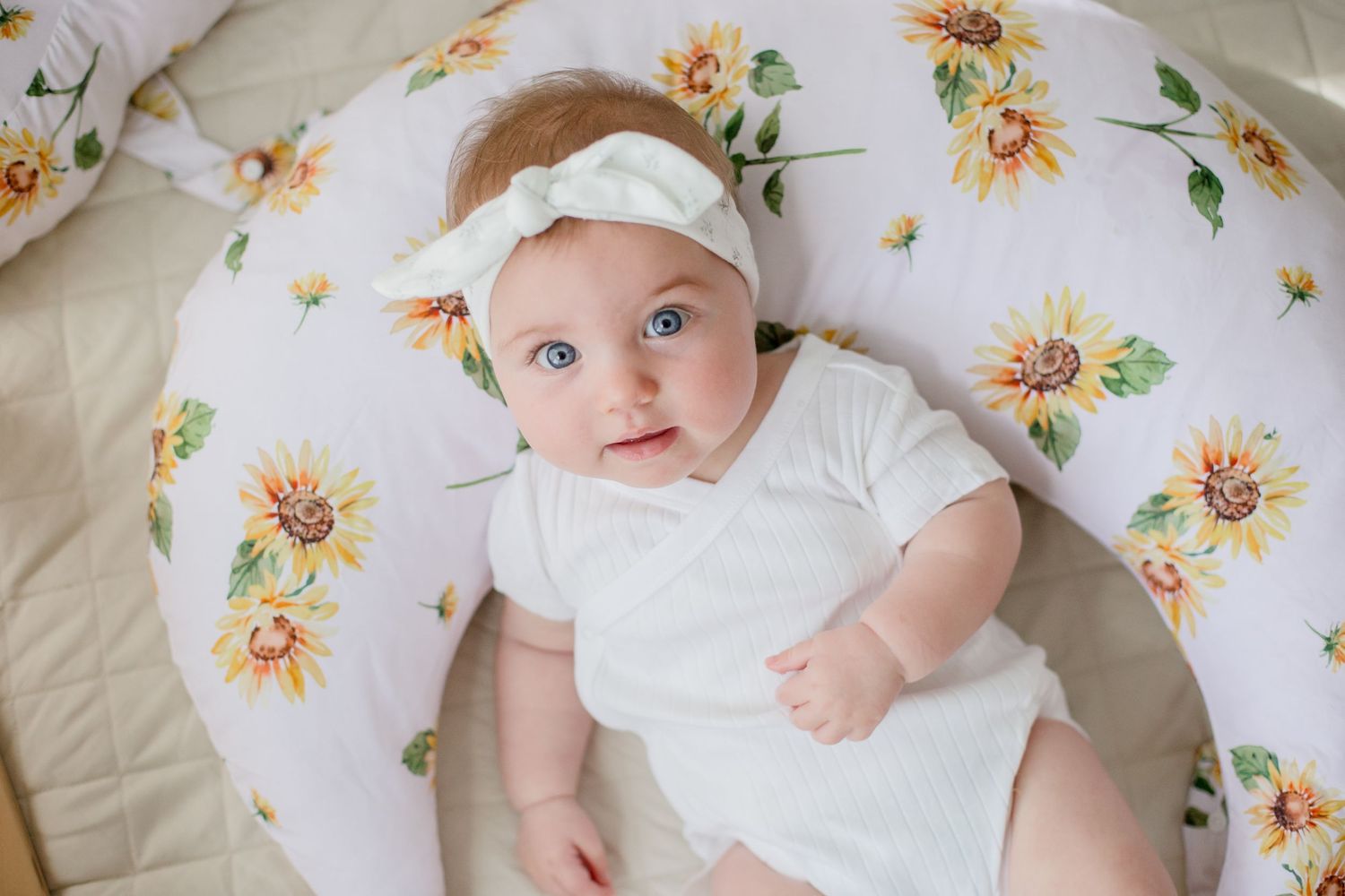 Baby in white onesie and headband lying on floral print bedding with sunflower pattern.