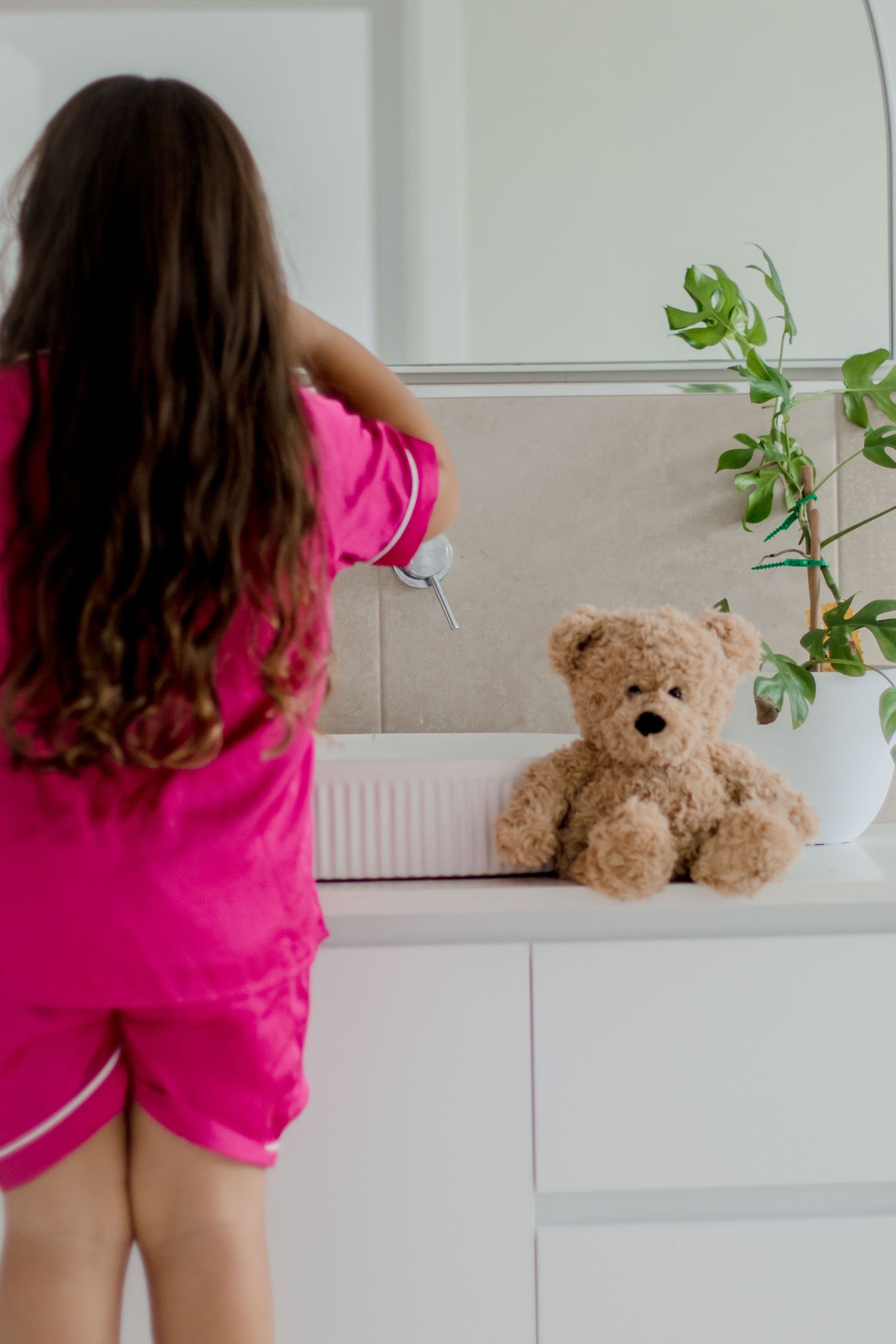 A child in pink pajamas looking at a teddy bear on a white shelf with plants.
