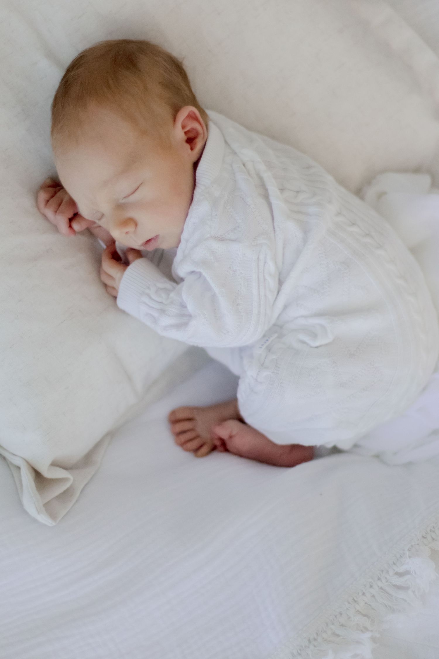 Sleeping baby in white onesie curled up on soft white bedding.