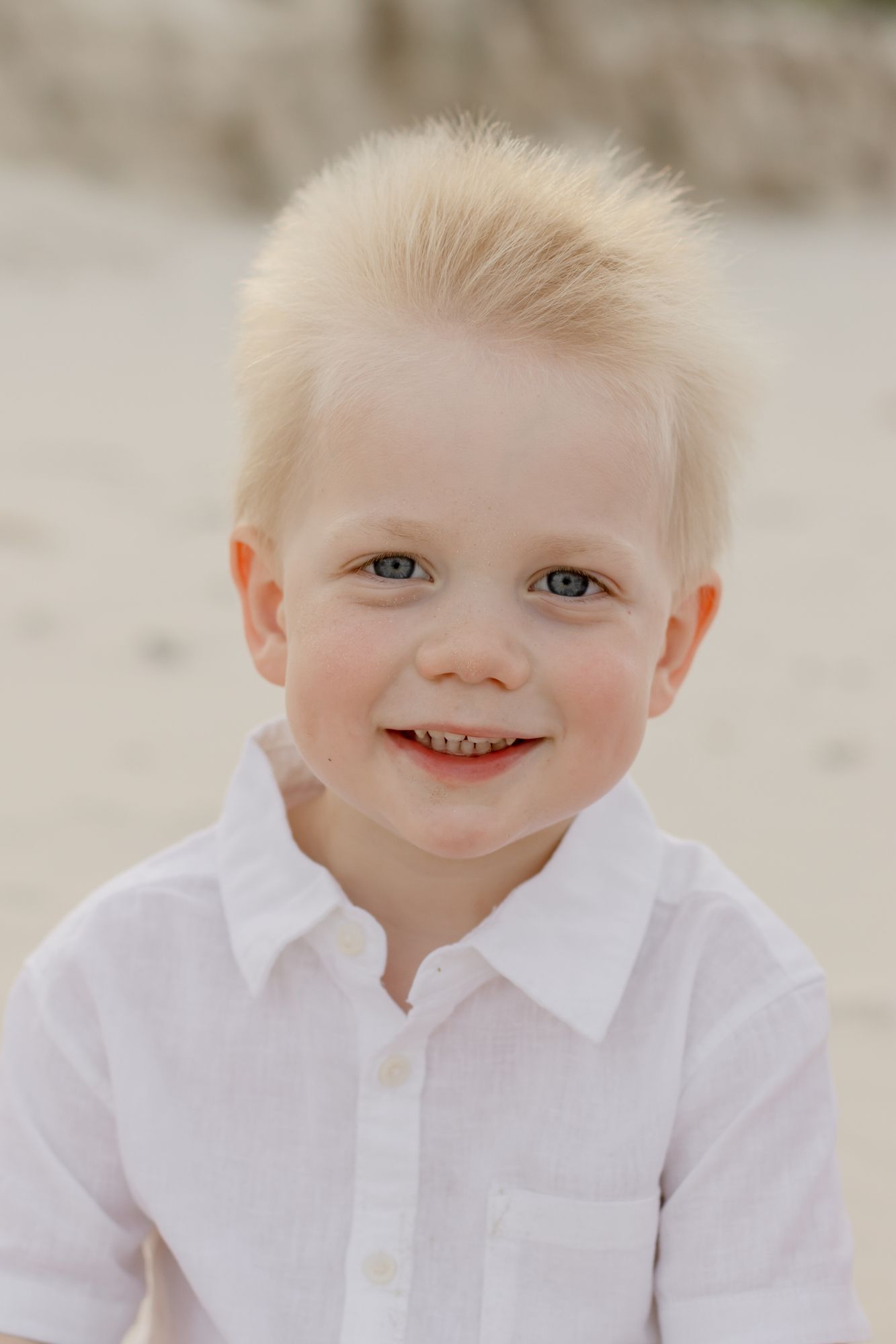 A young child in a white shirt smiles brightly at the camera on the beach.
