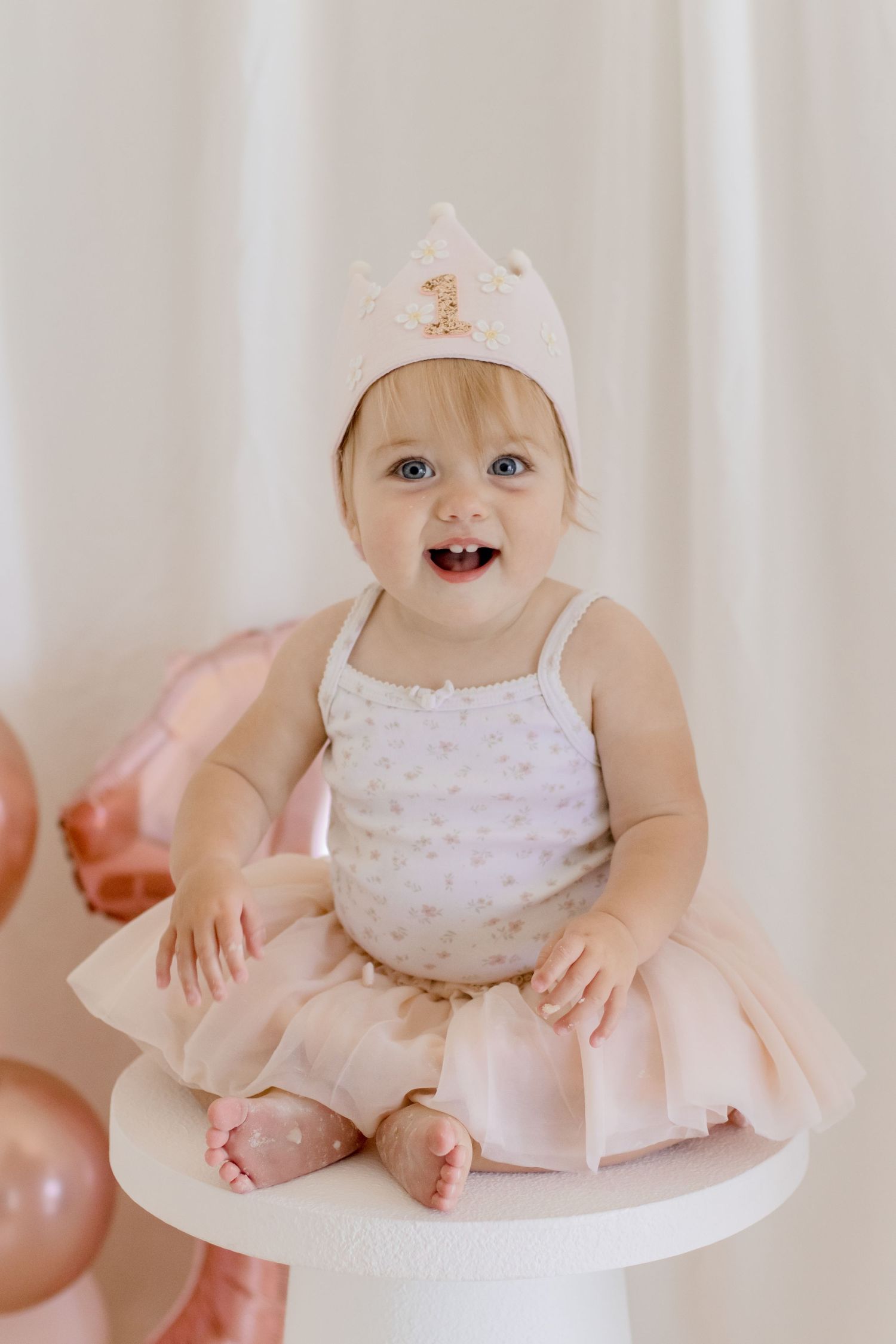 A joyful baby in a pink tutu and sparkly crown poses on a white table with pink balloons.