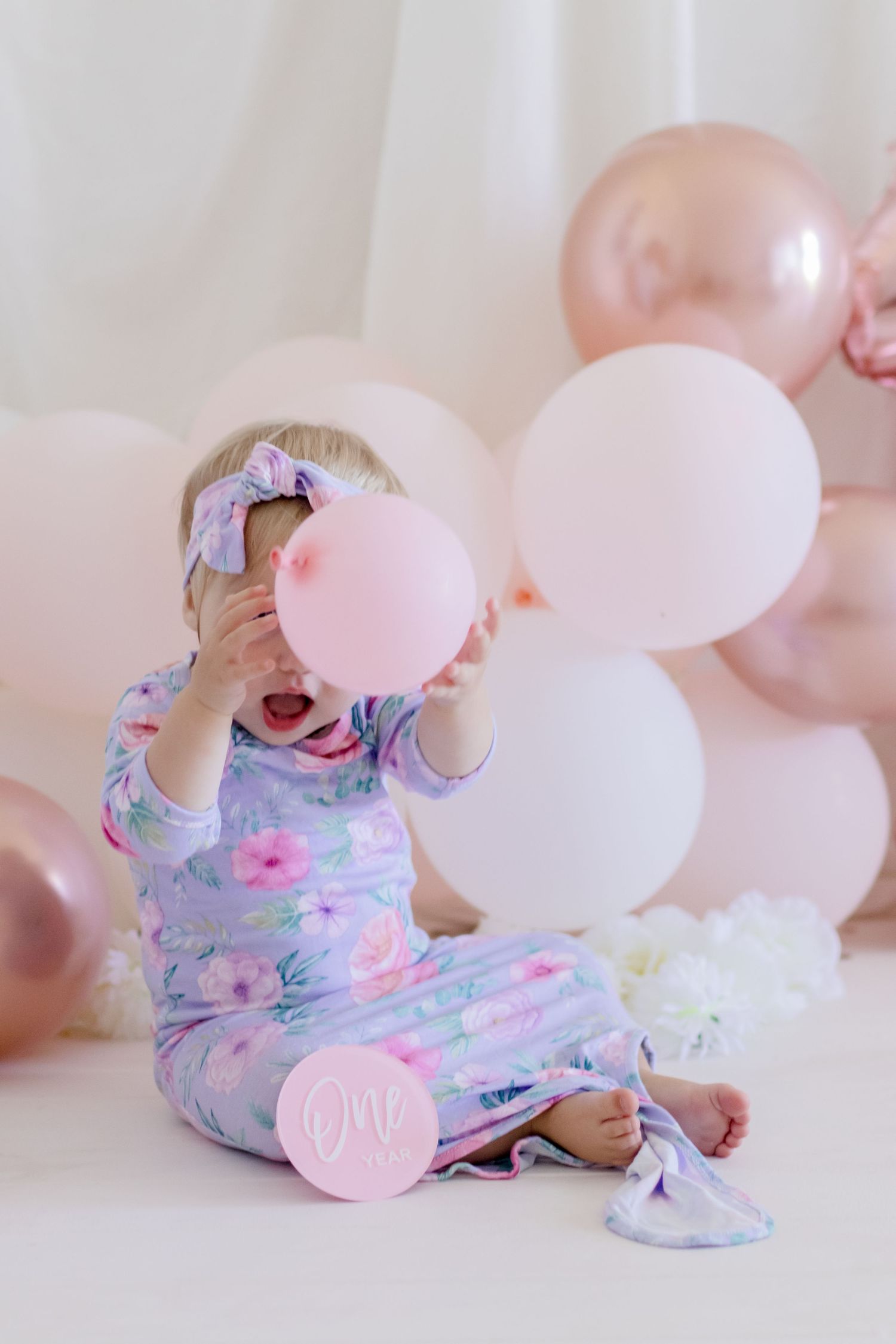 A floral pajama-clad infant plays with pink and white balloons during a photo session.