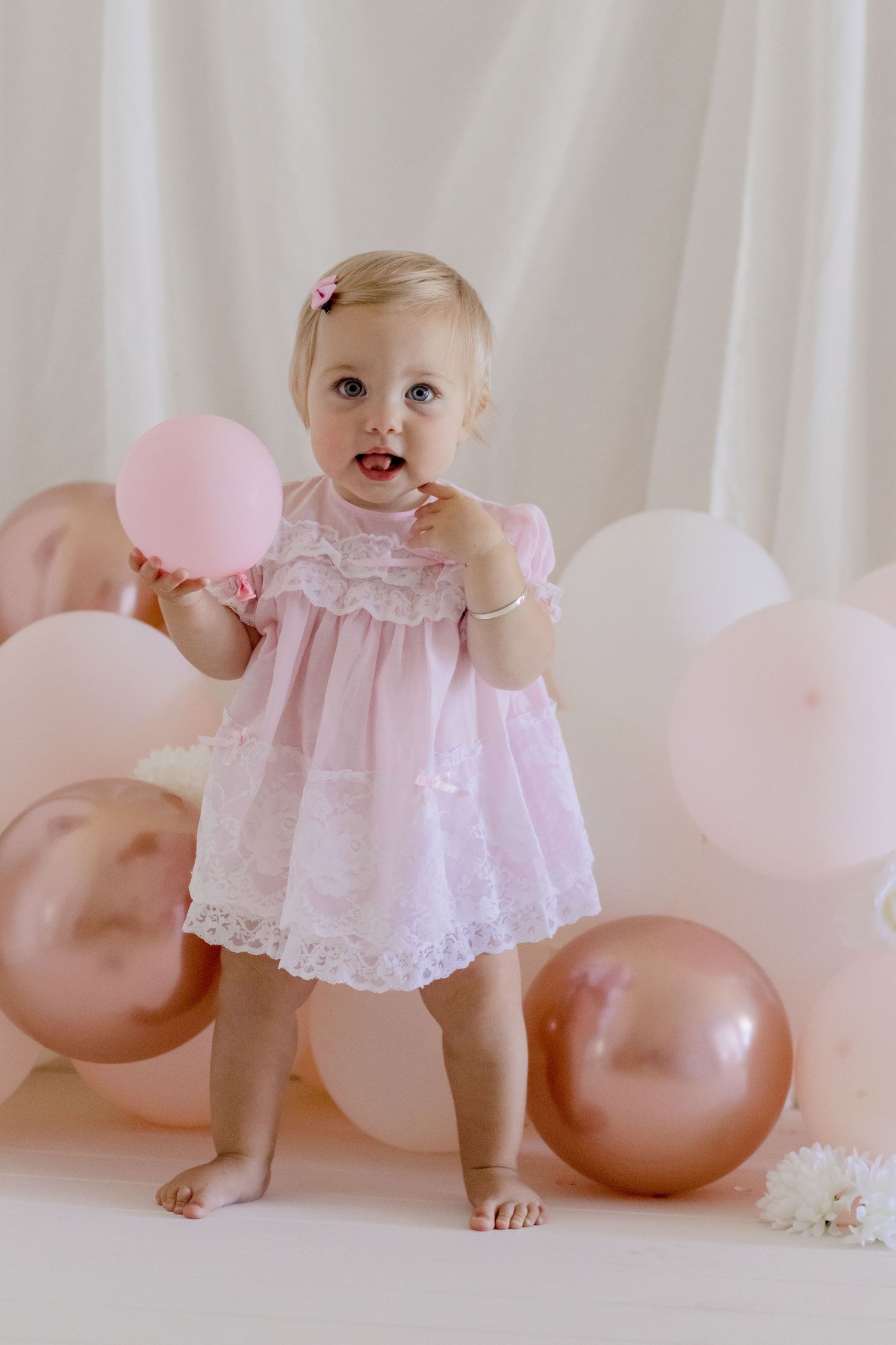 Happy baby holds pink balloons while wearing lace dress during photoshoot.