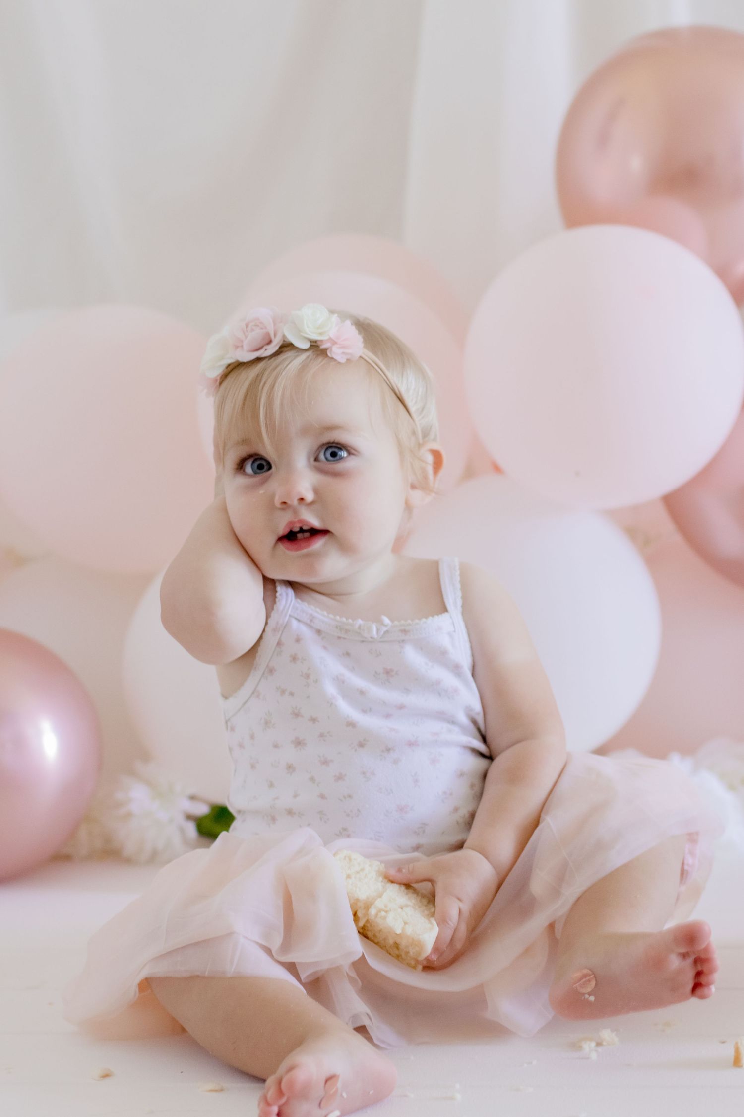 Baby with floral crown holding food while sitting against pink balloon background.