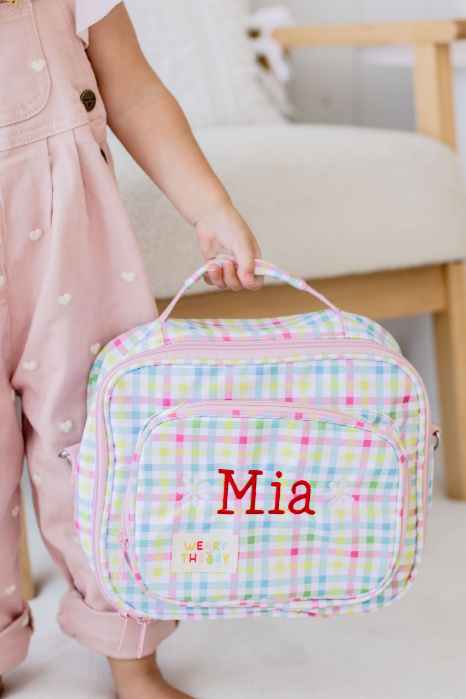 Girl holding a personalized pastel plaid lunch box with red embroidered name Mia, featuring rainbow checkered pattern.