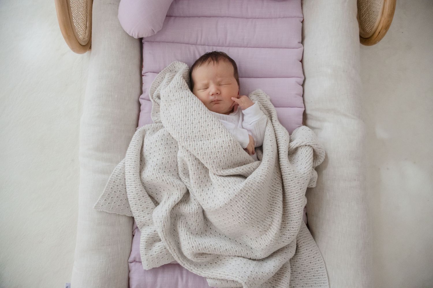 A sleeping newborn baby wrapped in a cozy beige knitted blanket rests on a lilac cushioned surface.
