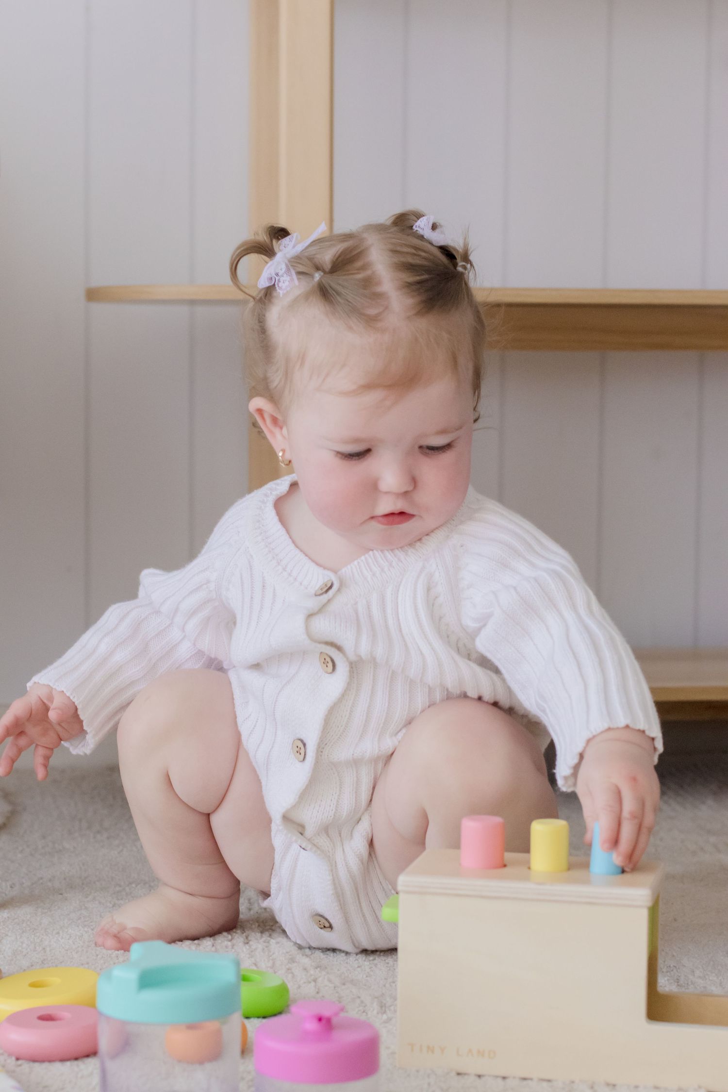 Small child in white romper playing with colorful wooden stacking toys on carpet.