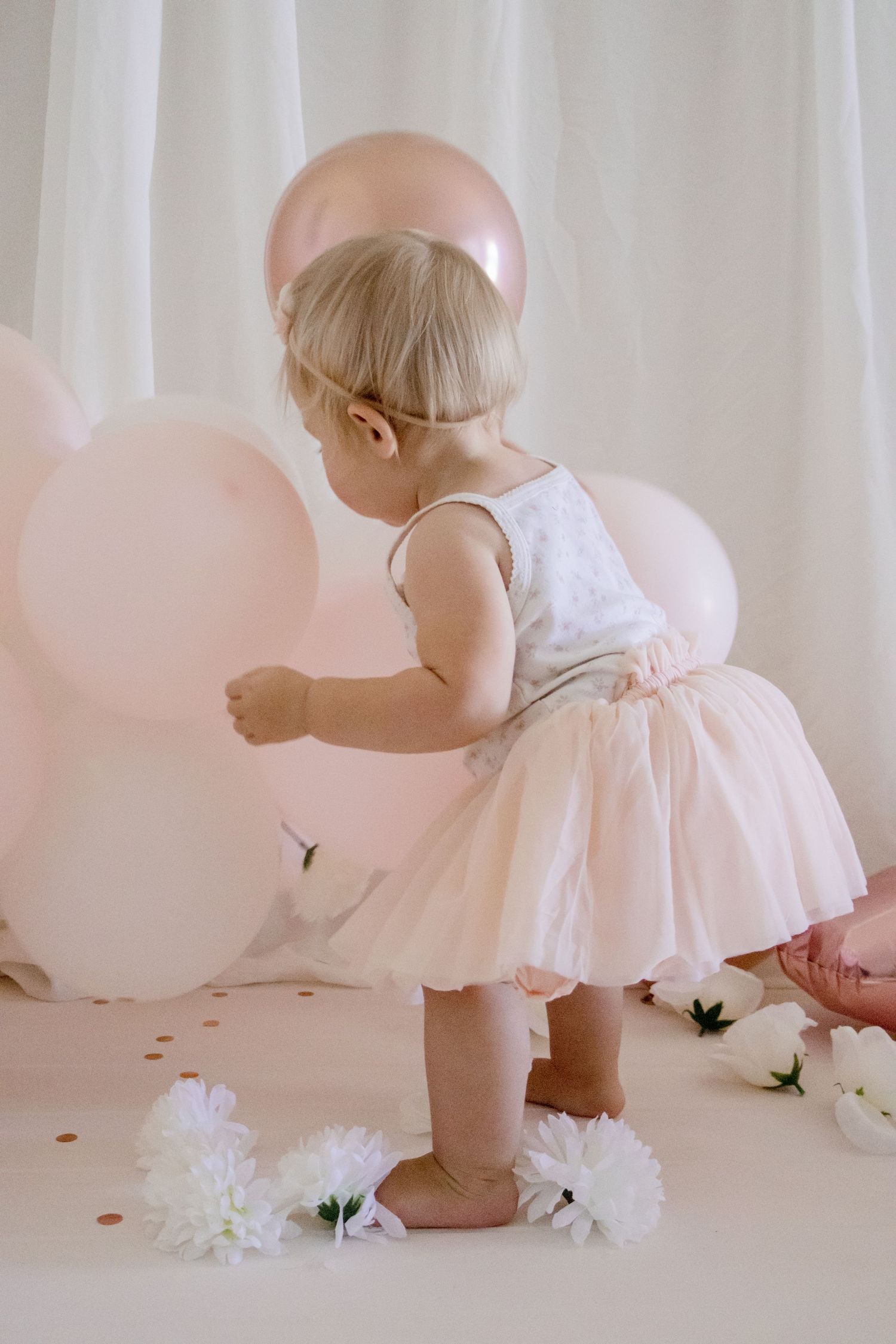 A small child in a white and pink dress explores festive balloons and flowers in a studio setting.