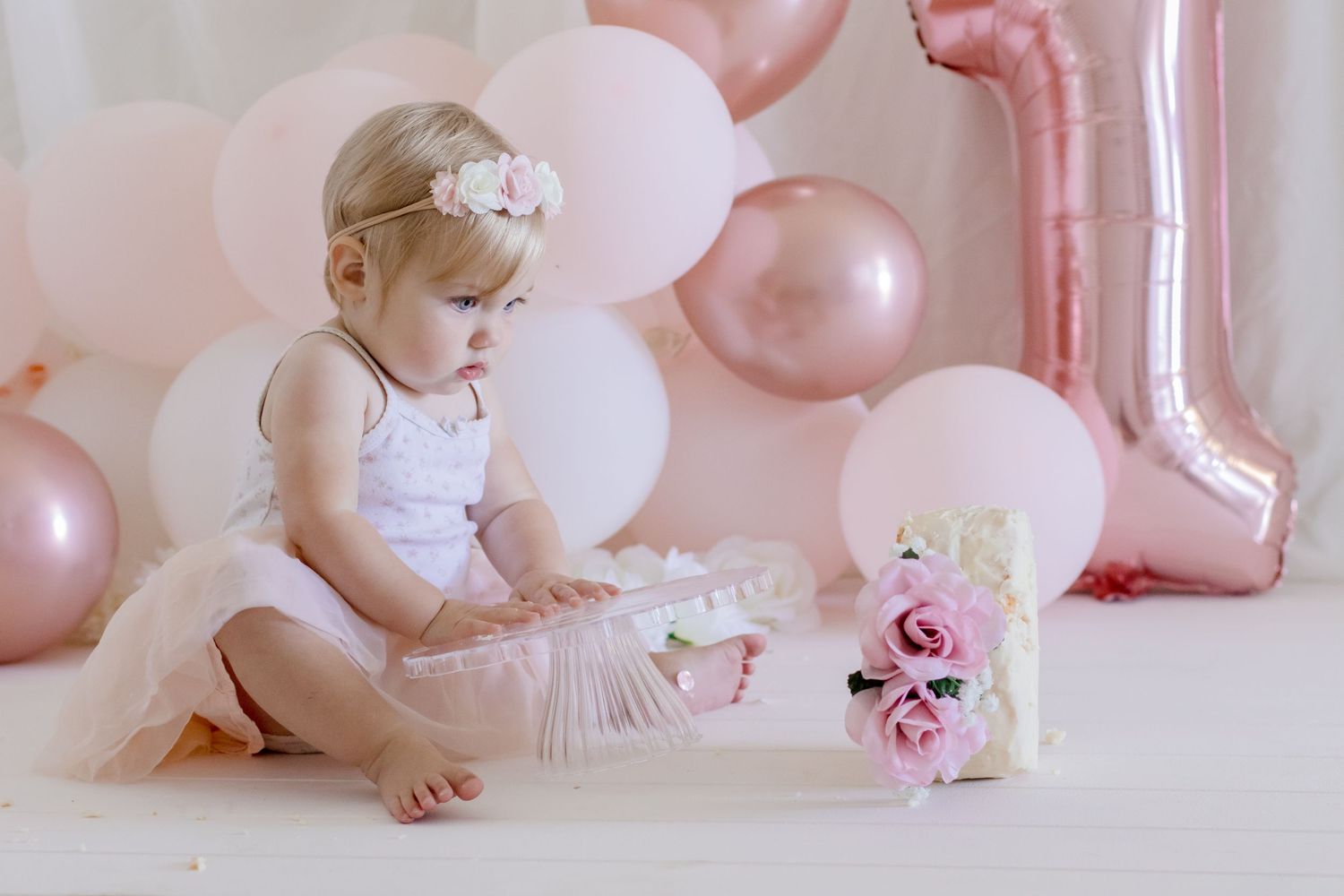 Baby in white tulle dress exploring birthday decorations with pink roses and balloons.