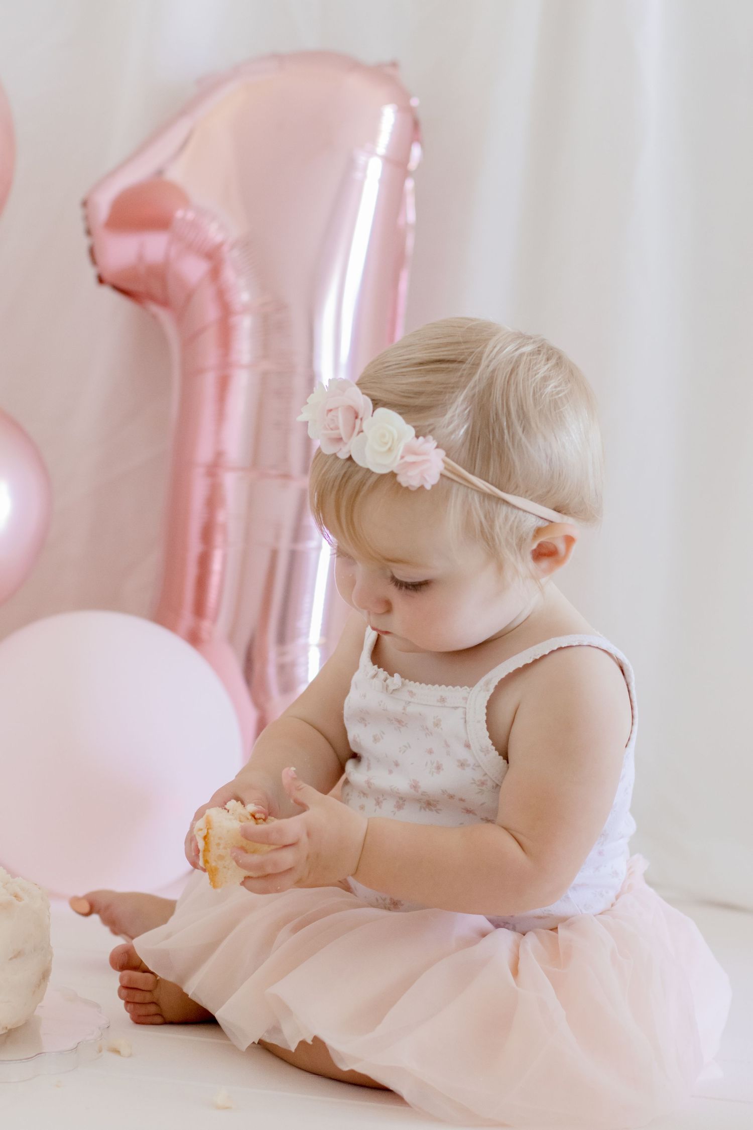 Baby in white dress examining snack with pink number one balloon in background.