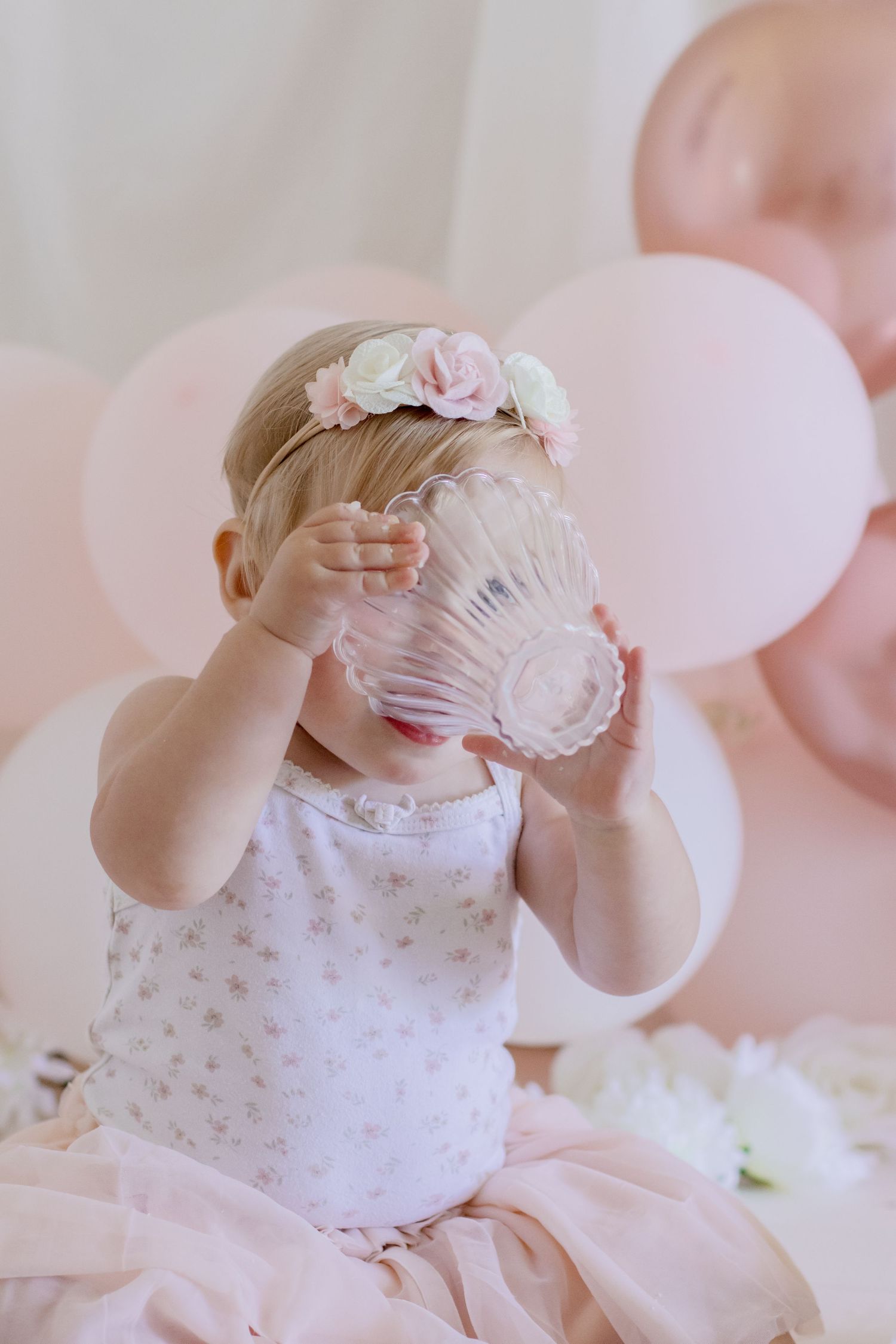 Baby in white dress with floral headband drinking from crystal glass against pink balloon backdrop.