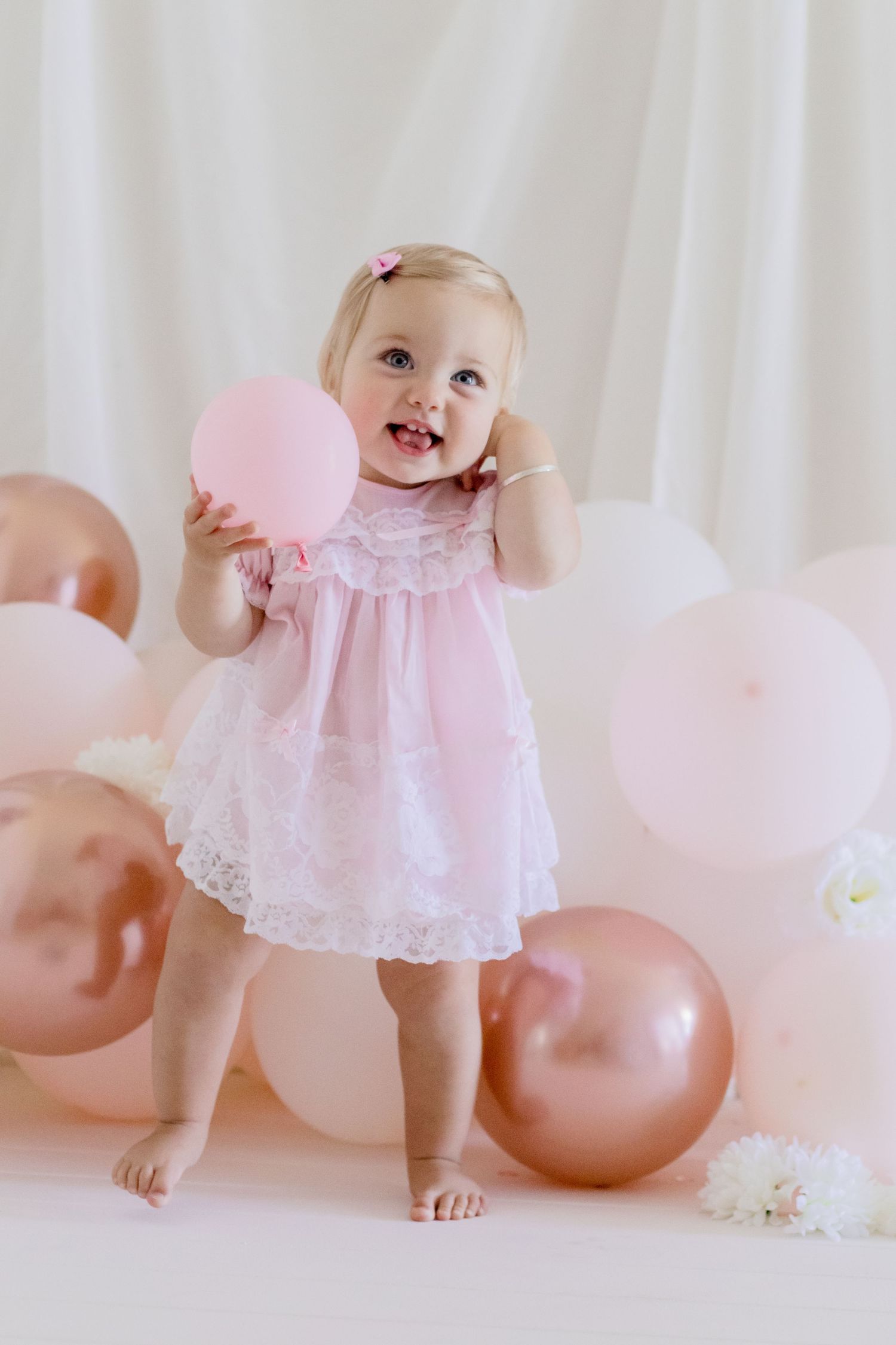 Joyful baby wearing pink lace dress poses with pink balloons in studio setting.