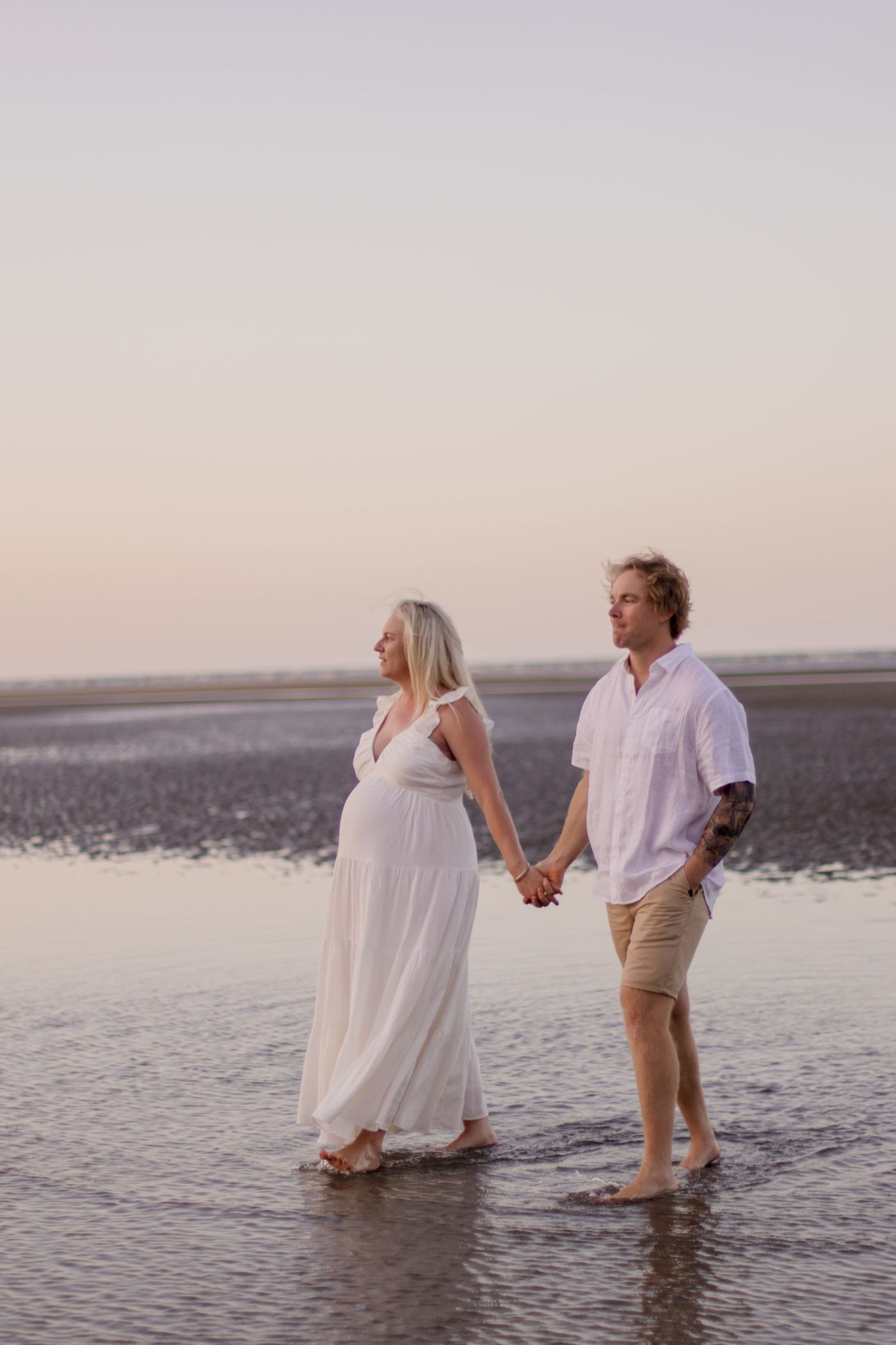 A couple walks hand in hand through shallow water during their beach maternity session.