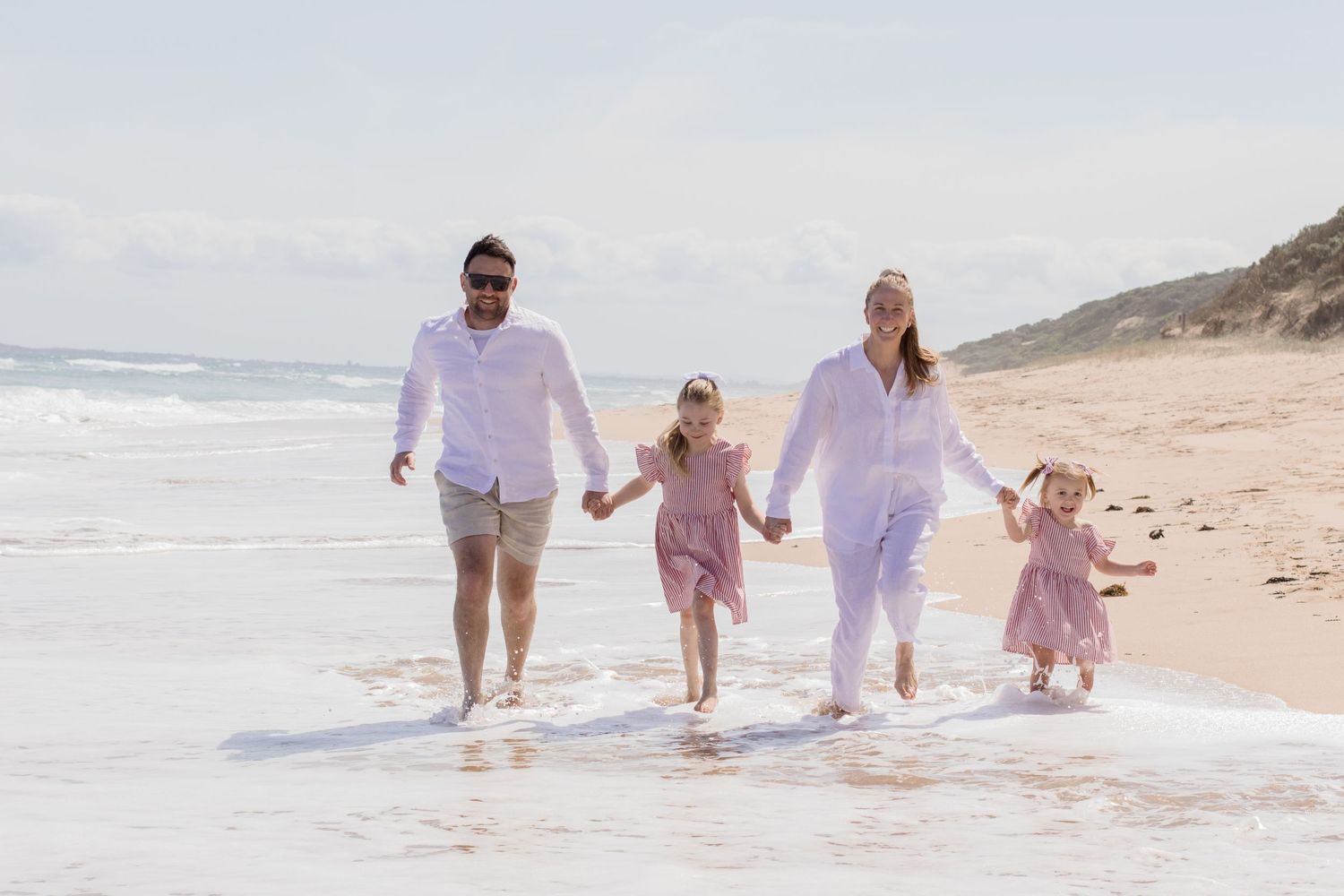 Group holding hands while walking together along the shoreline in matching light colored outfits.