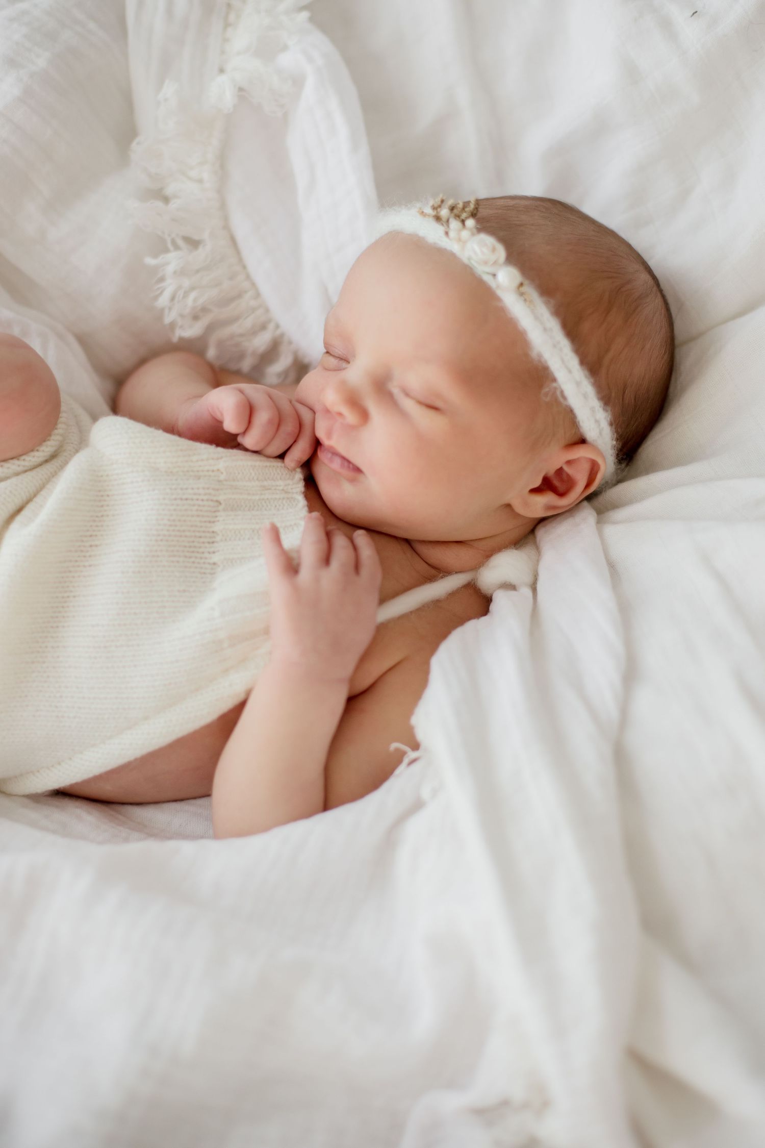 Sleeping newborn baby wearing white lace headband lying on white bedding.