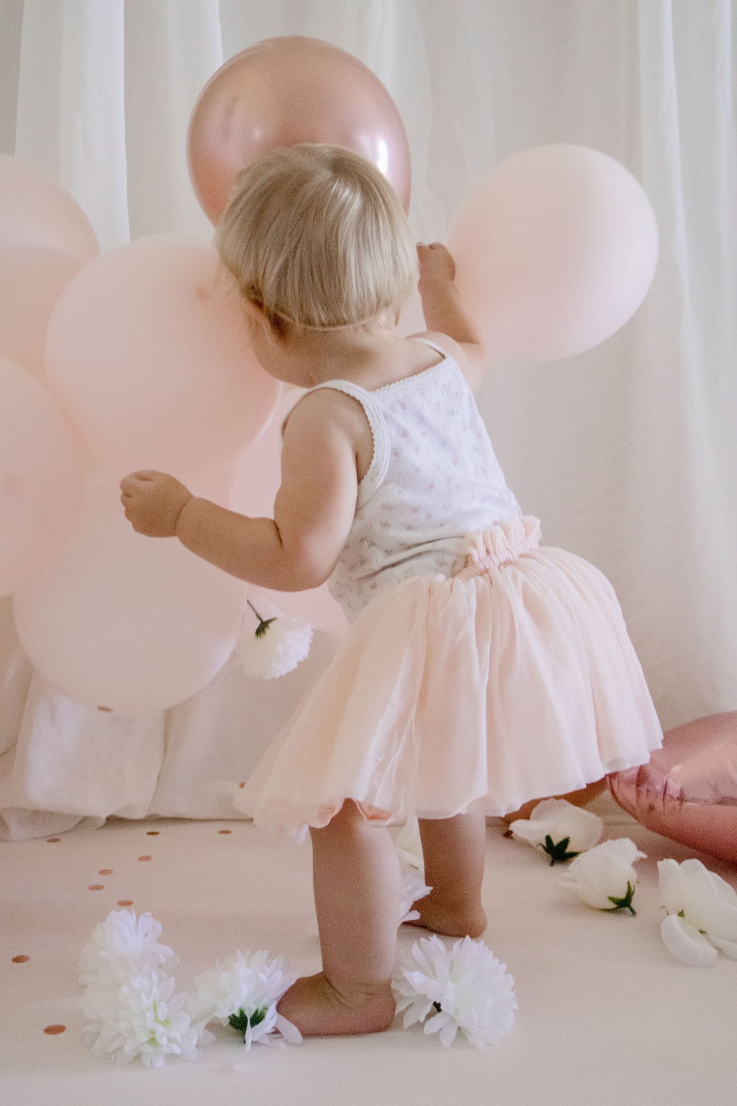 A young child in a party dress plays with pink and rose gold balloons against white drapery.