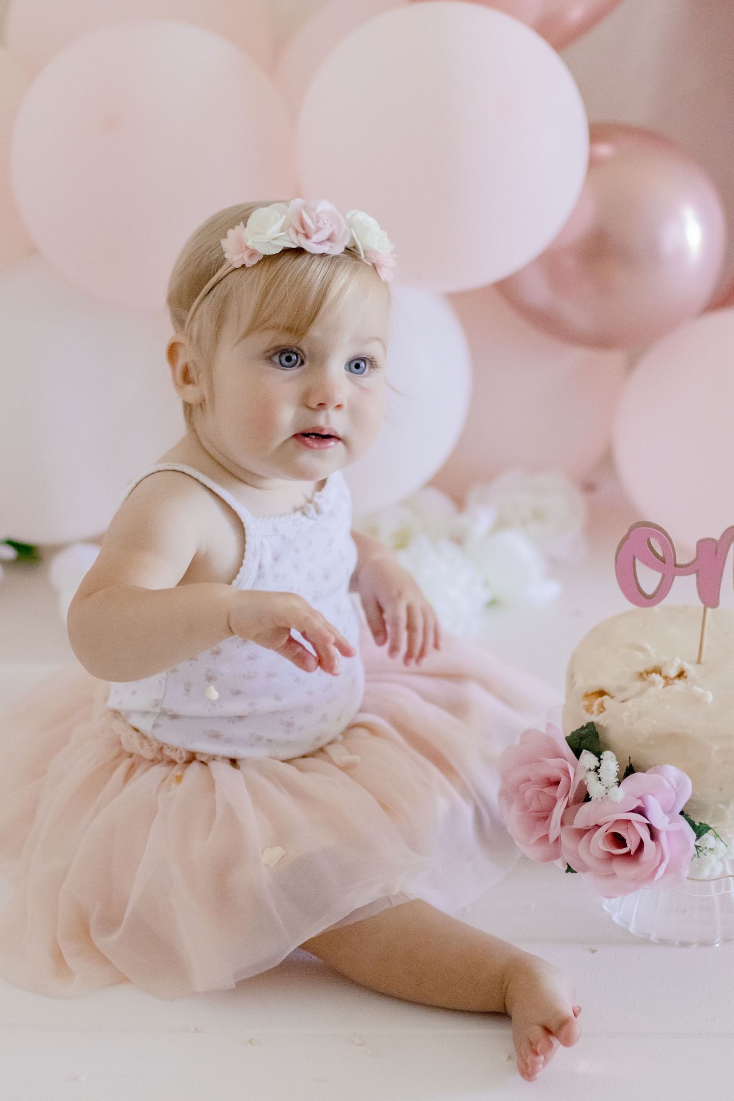 Baby in tulle dress and floral crown at first birthday photoshoot with pink balloons.