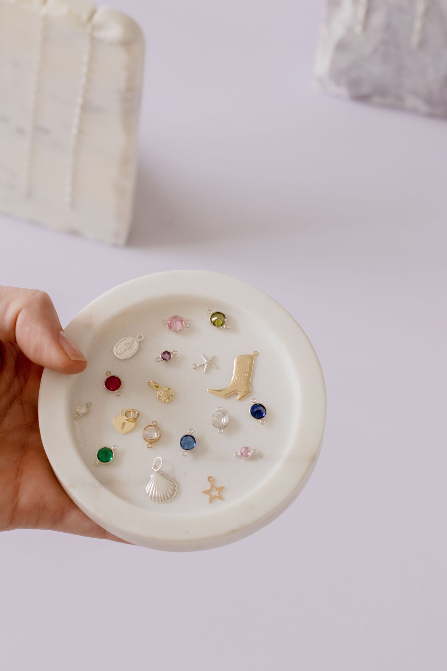 Hand holding white dish filled with small earring studs in various colors and dainty charm designs.