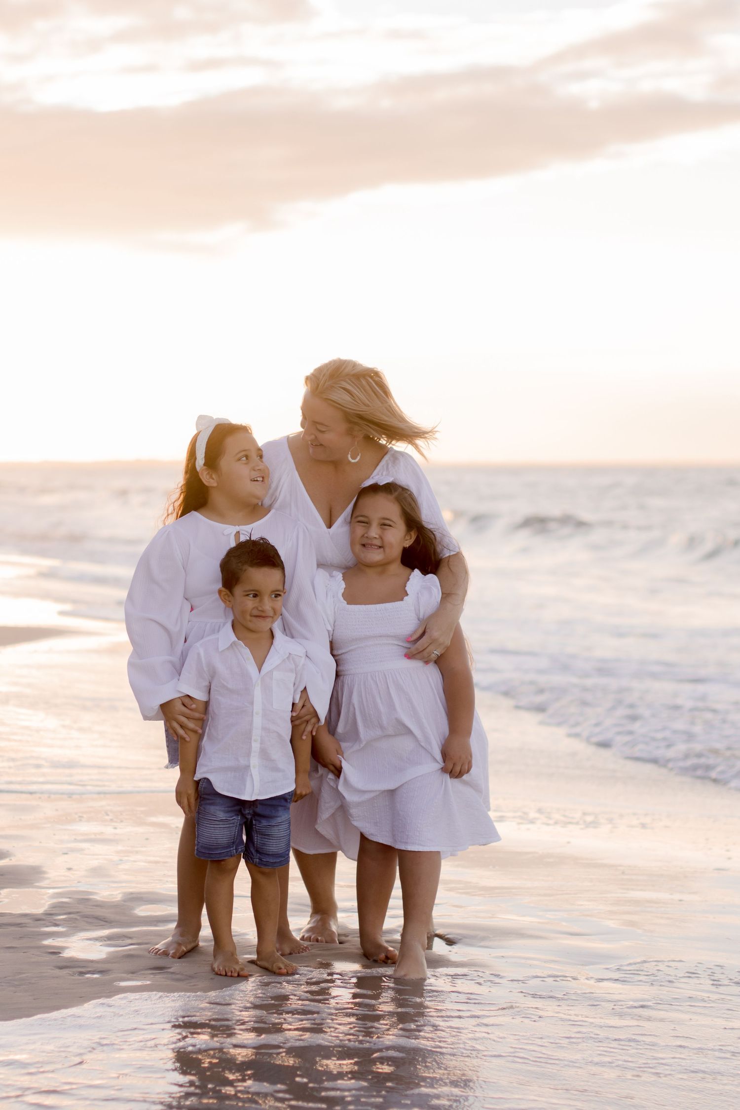 A family wearing white outfits plays together at the beach during sunset as waves lap at their feet.