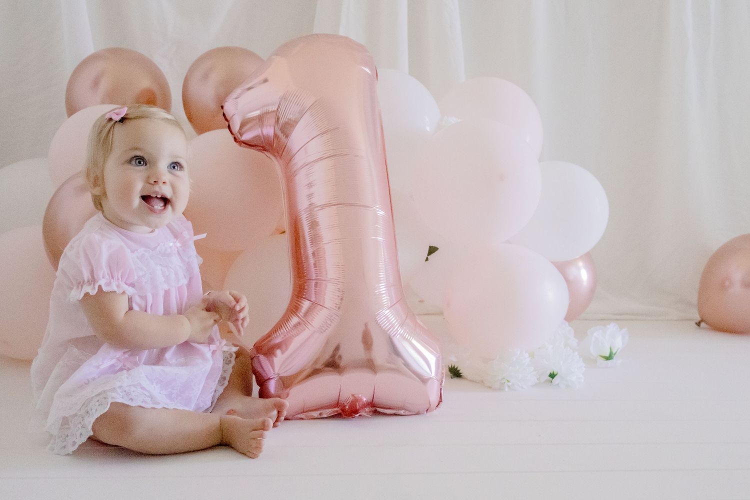 Happy baby in pink dress sitting next to rose gold number one balloon with white background.