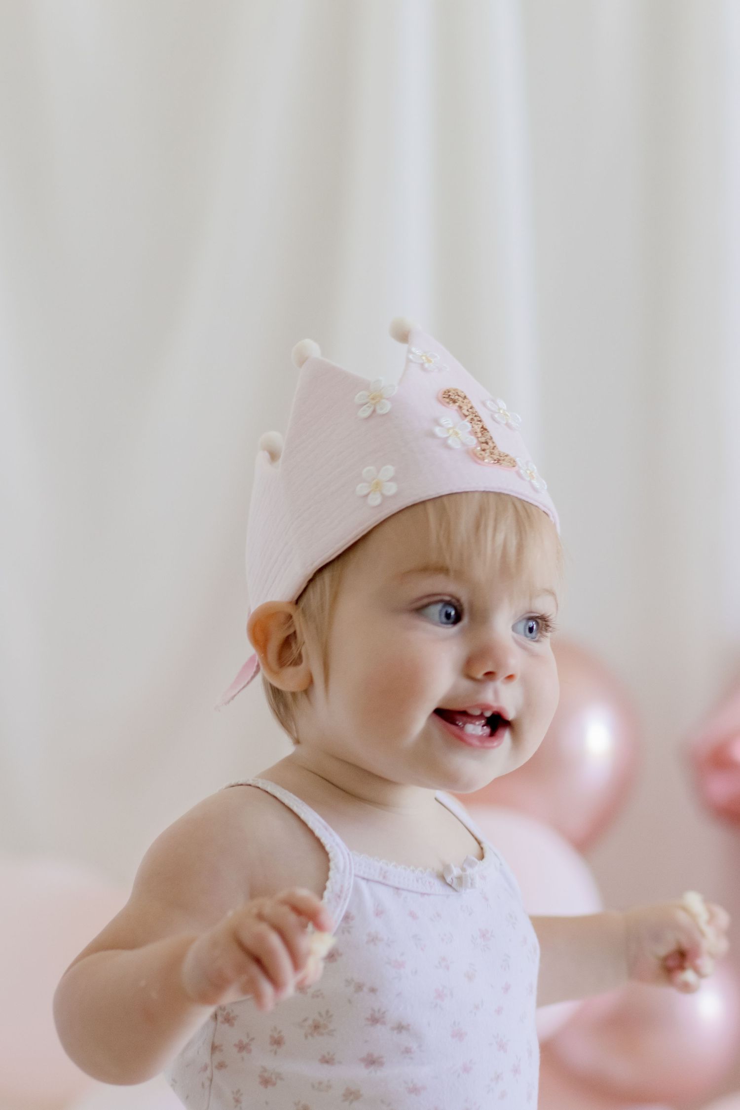 A joyful toddler in white dress wearing a decorative crown with pink balloons in background.