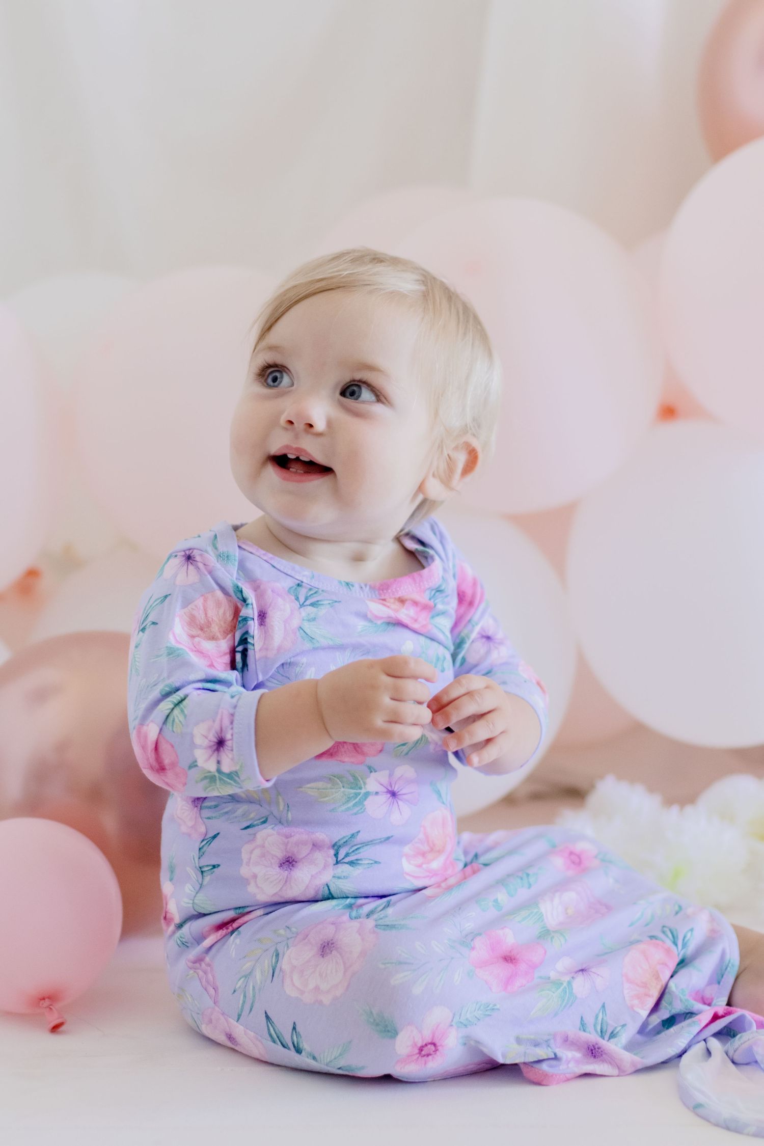 Baby in floral sleepwear looking up joyfully during pink balloon photoshoot.