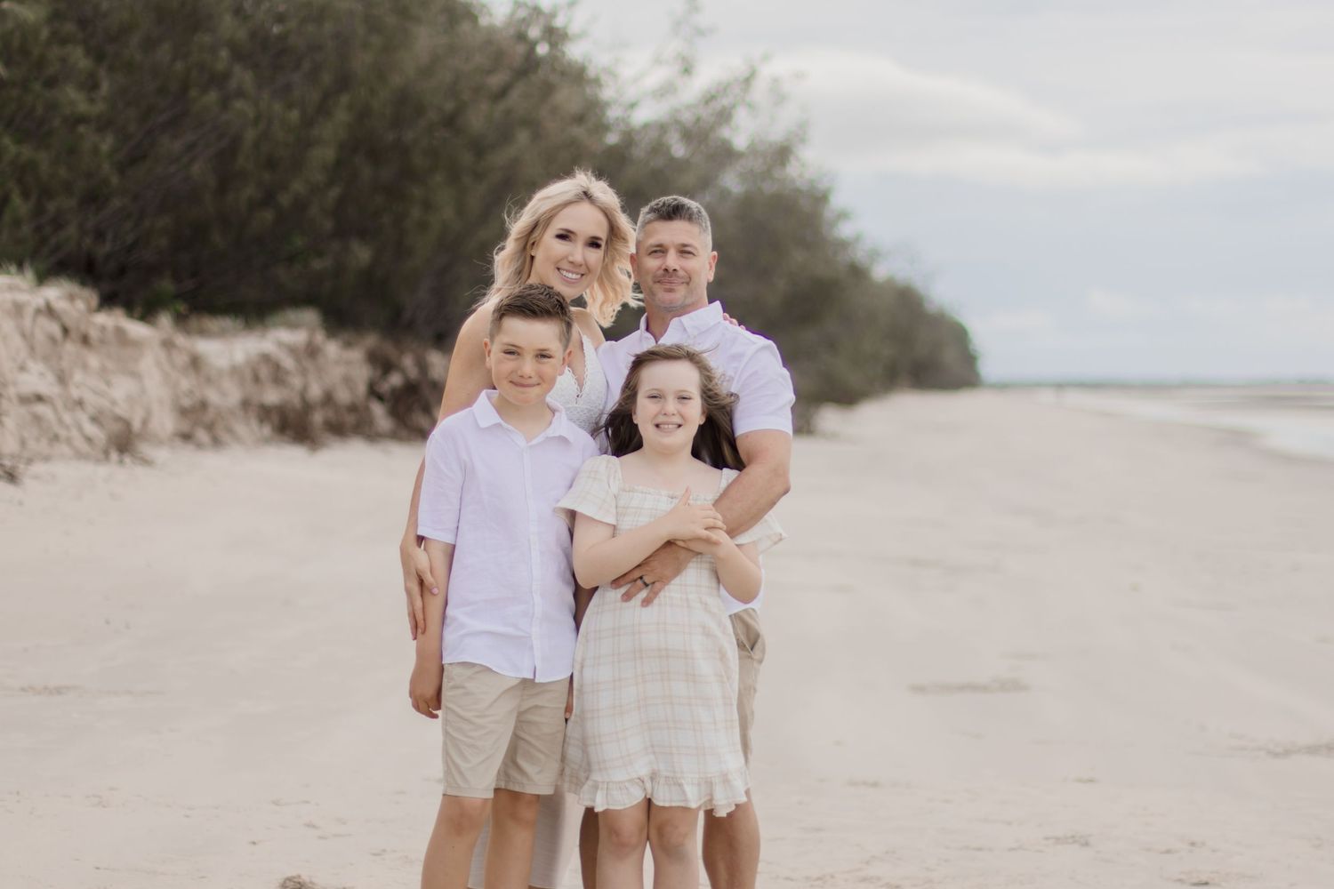 Happy family posing together for a portrait on a breezy beach with trees in background.