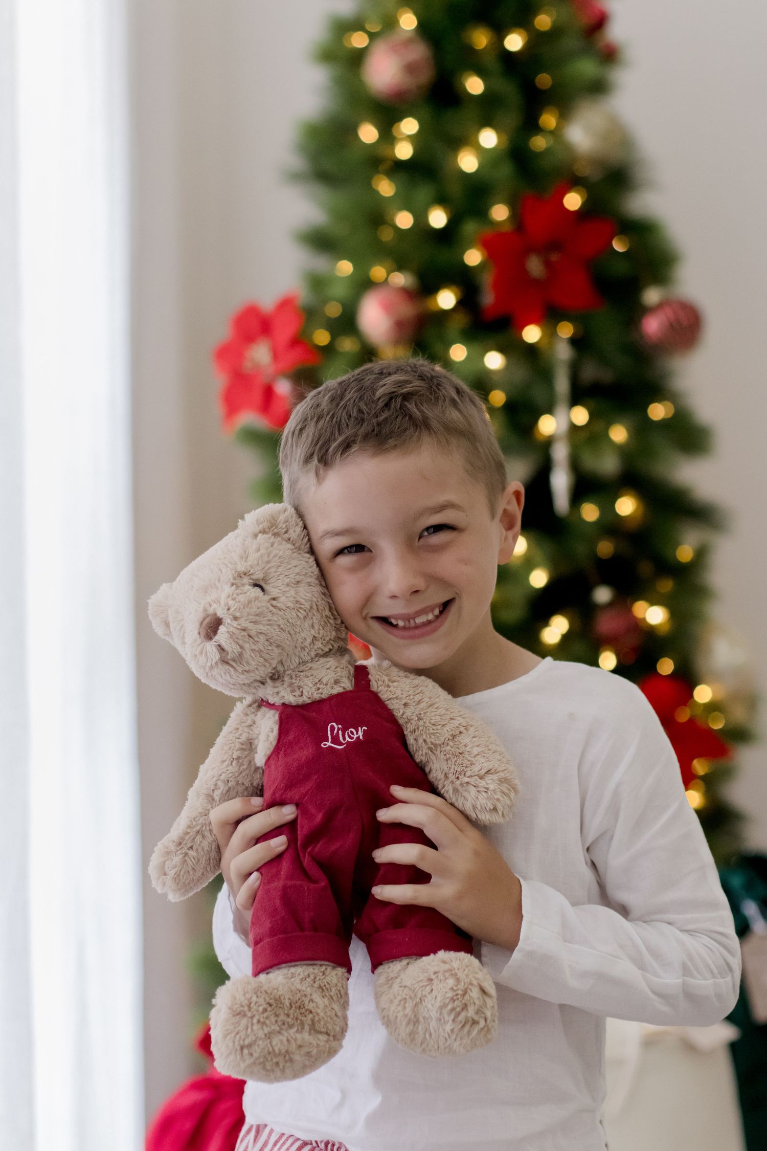 A child in a white shirt holds a plush teddy bear wearing a red sweater in front of a decorated Christmas tree.