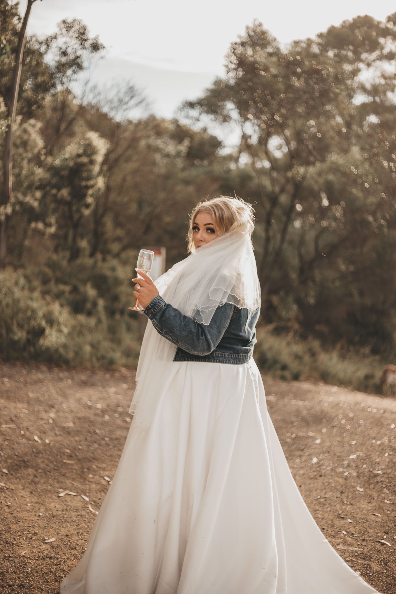 A romantic embrace between figures in wedding attire with a flowing white dress and denim jacket against a natural backdrop.