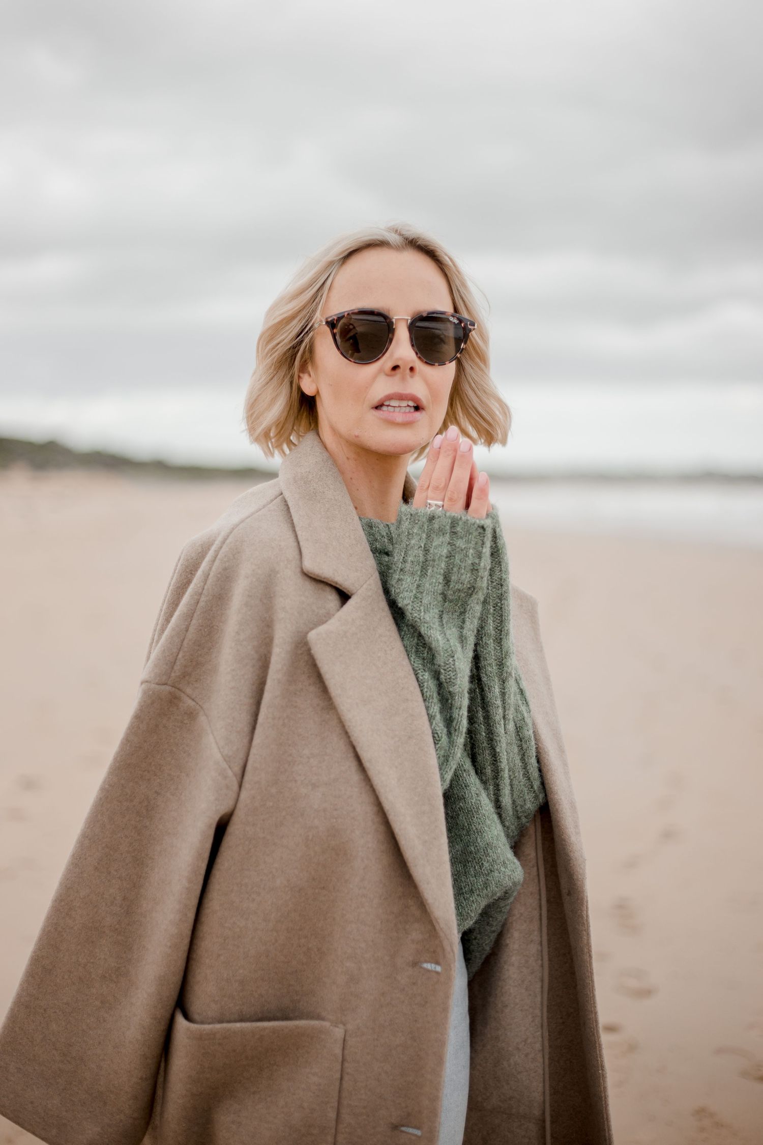 Fashionable beige overcoat and green sweater worn at cloudy beach with sunglasses and casual styling.